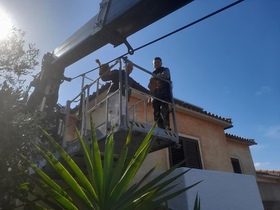 Due operai su una piattaforma elevatrice, che installano qualcosa vicino a una casa. Sole splendente e cielo azzurro.