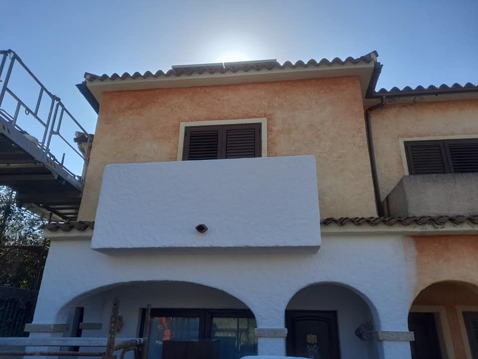 Edificio in stucco a due piani con balcone bianco e ingresso ad arco al piano terra, sotto un sole splendente.