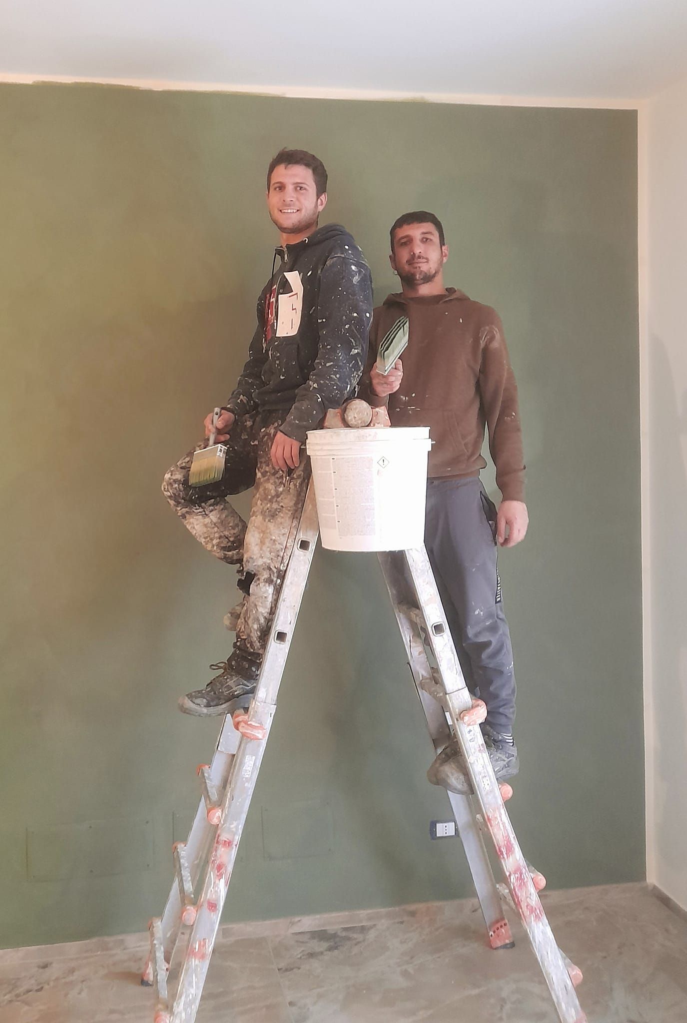 Due uomini stanno dipingendo un muro. Uno è su una scala con un secchio di vernice, l'altro è in piedi accanto.