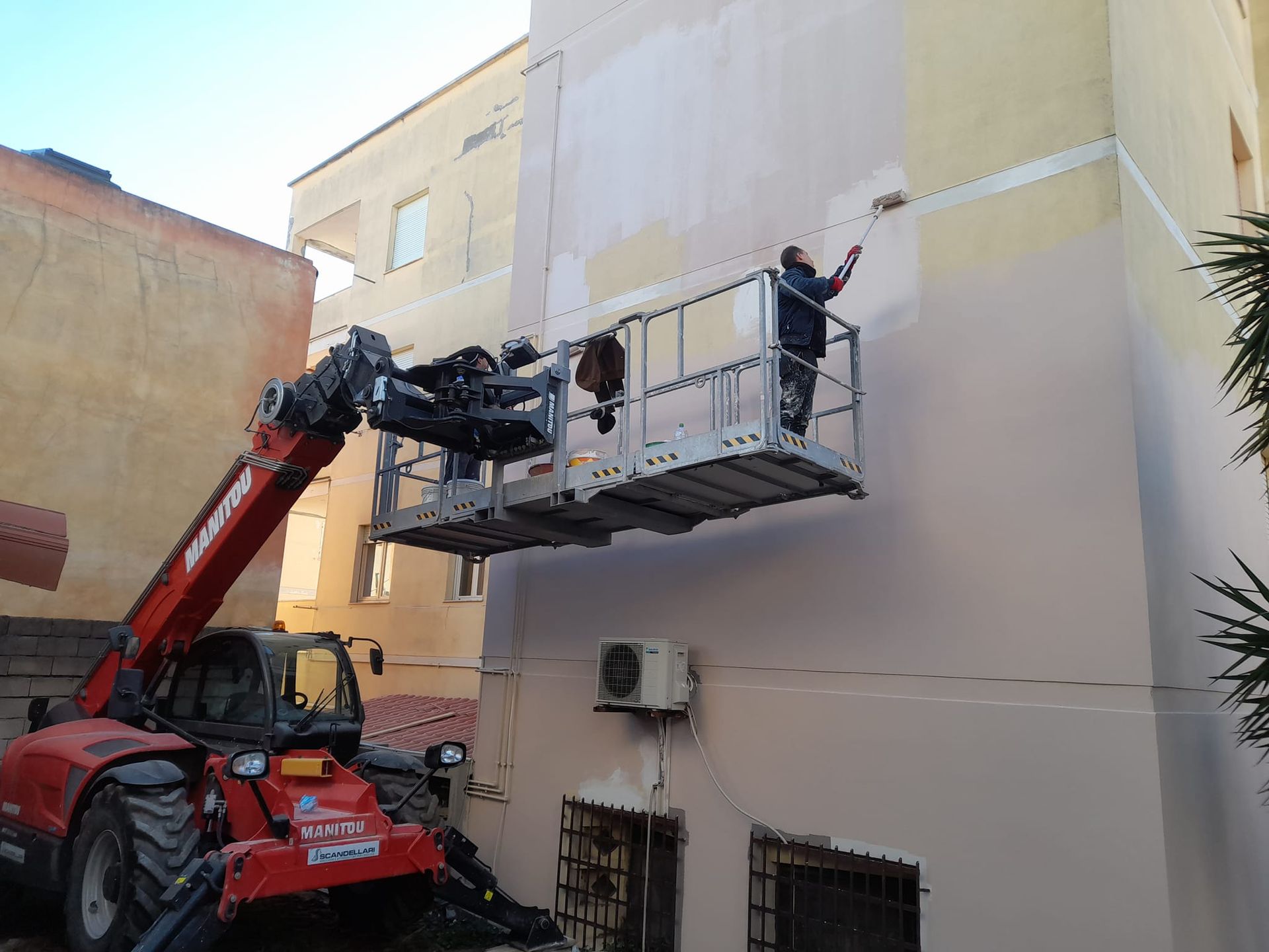 Persona che dipinge la facciata di un edificio da una piattaforma elevatrice sollevata da un sollevatore telescopico rosso.