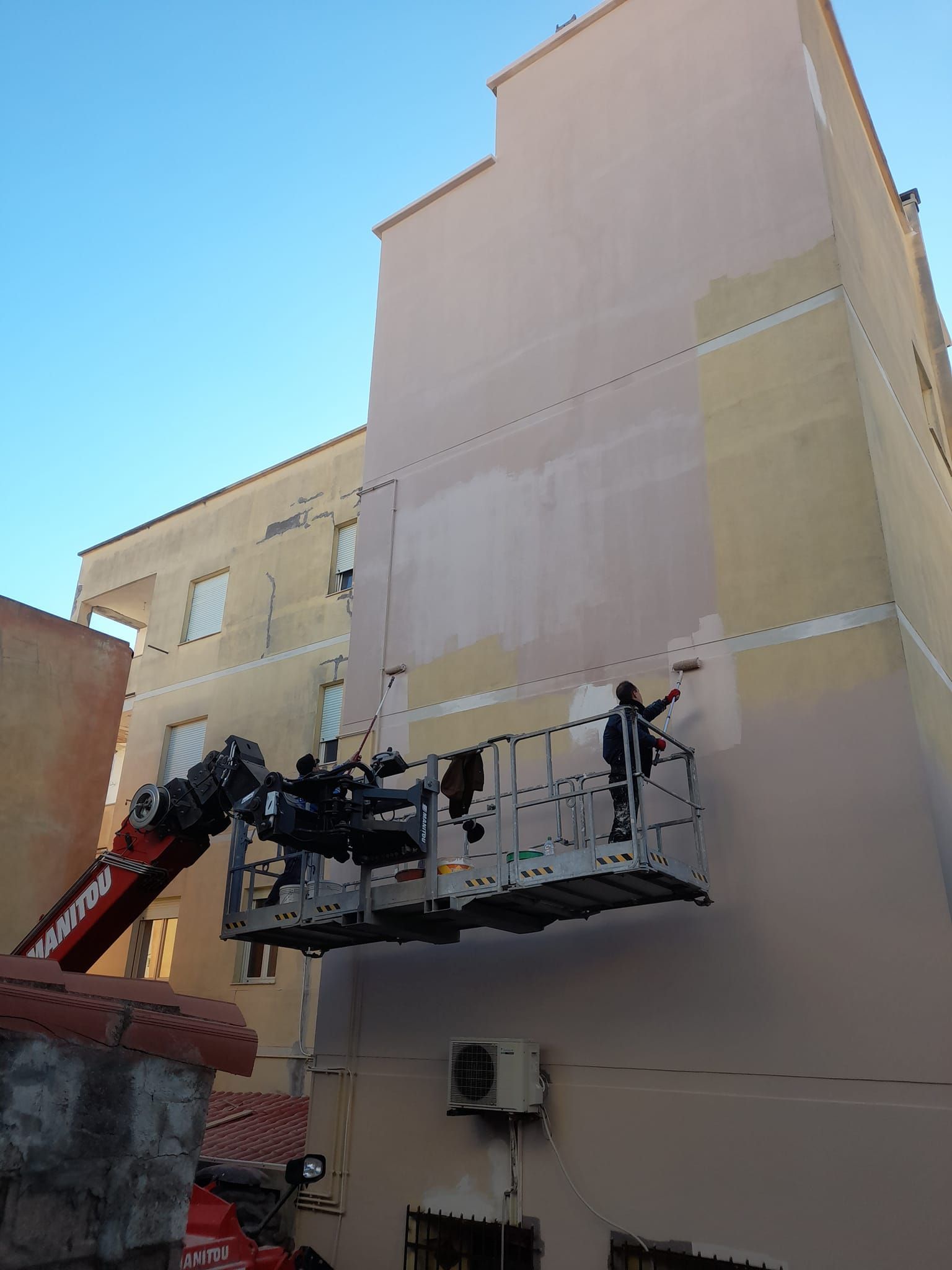 Operai in ascensore che dipingono l'esterno di un edificio di colore chiaro, sotto un cielo azzurro e terso.