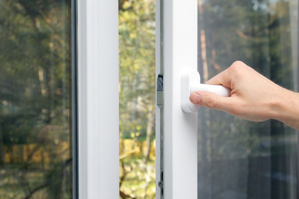 Hand opening a white window with a view of green trees outside.
