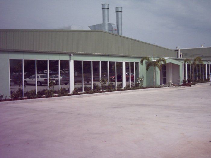 Exterior of a light green industrial building with a long row of windows, palm trees, and smokestacks in the background.