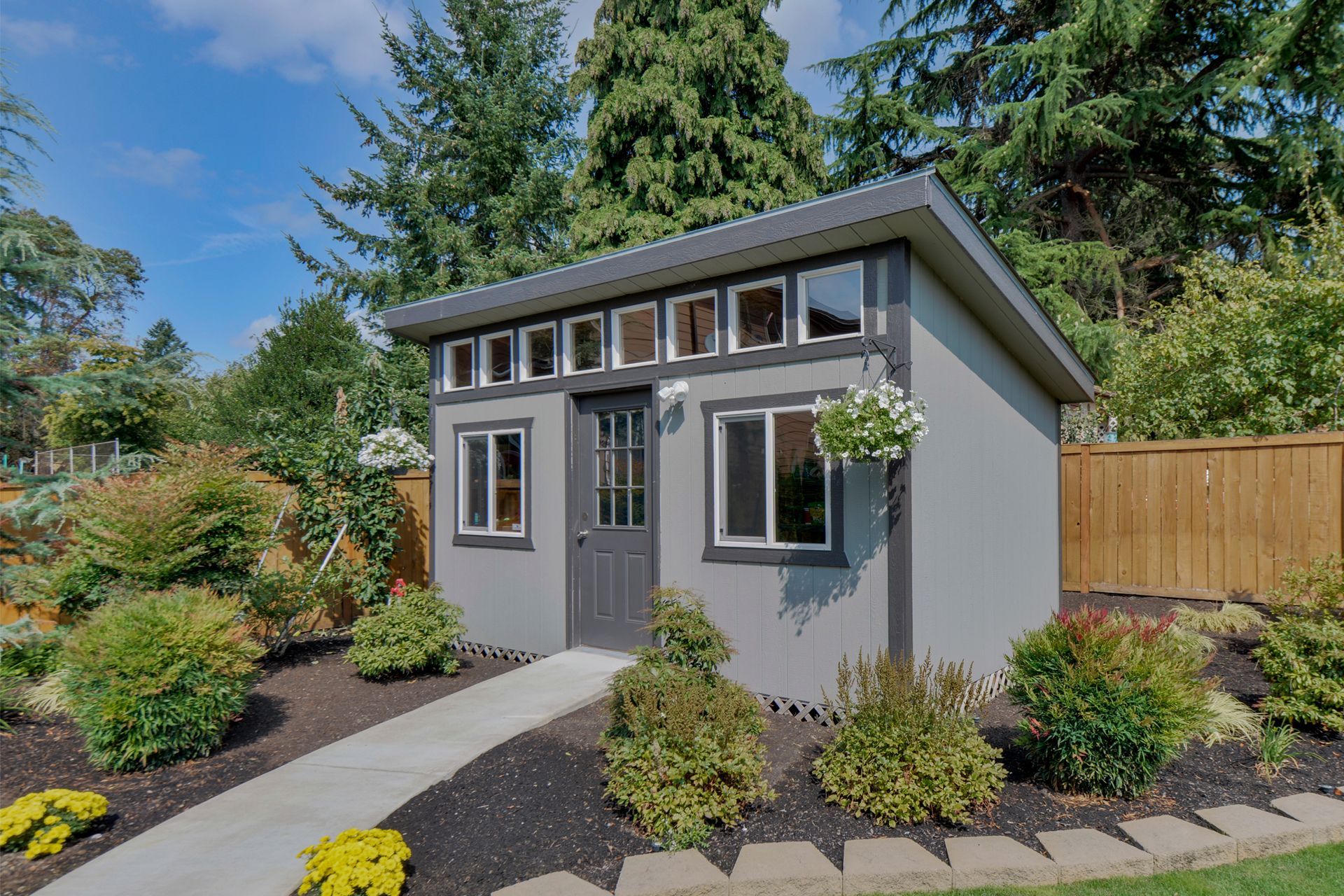 Grey shed with windows, a door, and a walkway, surrounded by landscaping and trees.