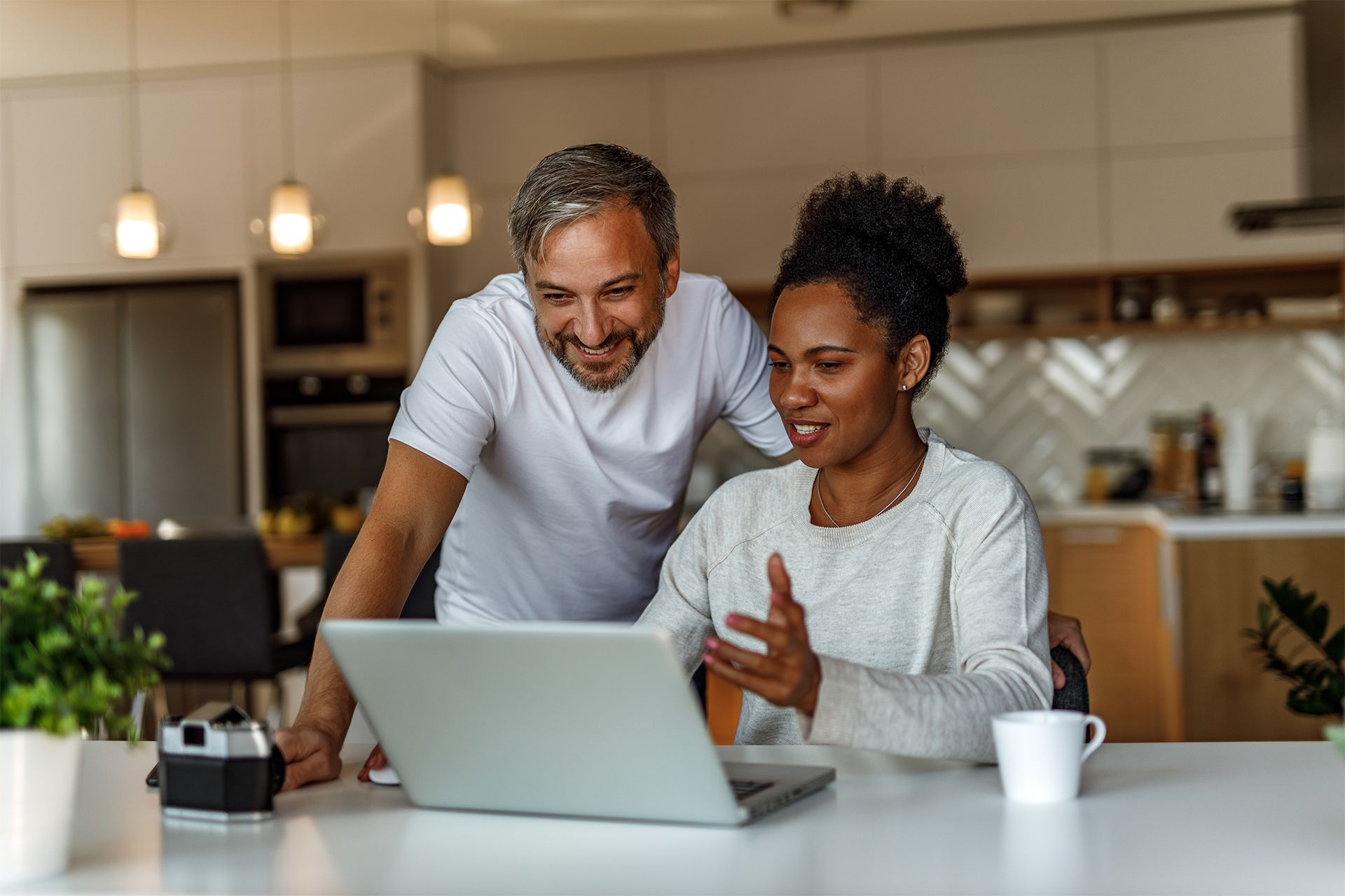 A couple smiles at a laptop screen in a bright kitchen.