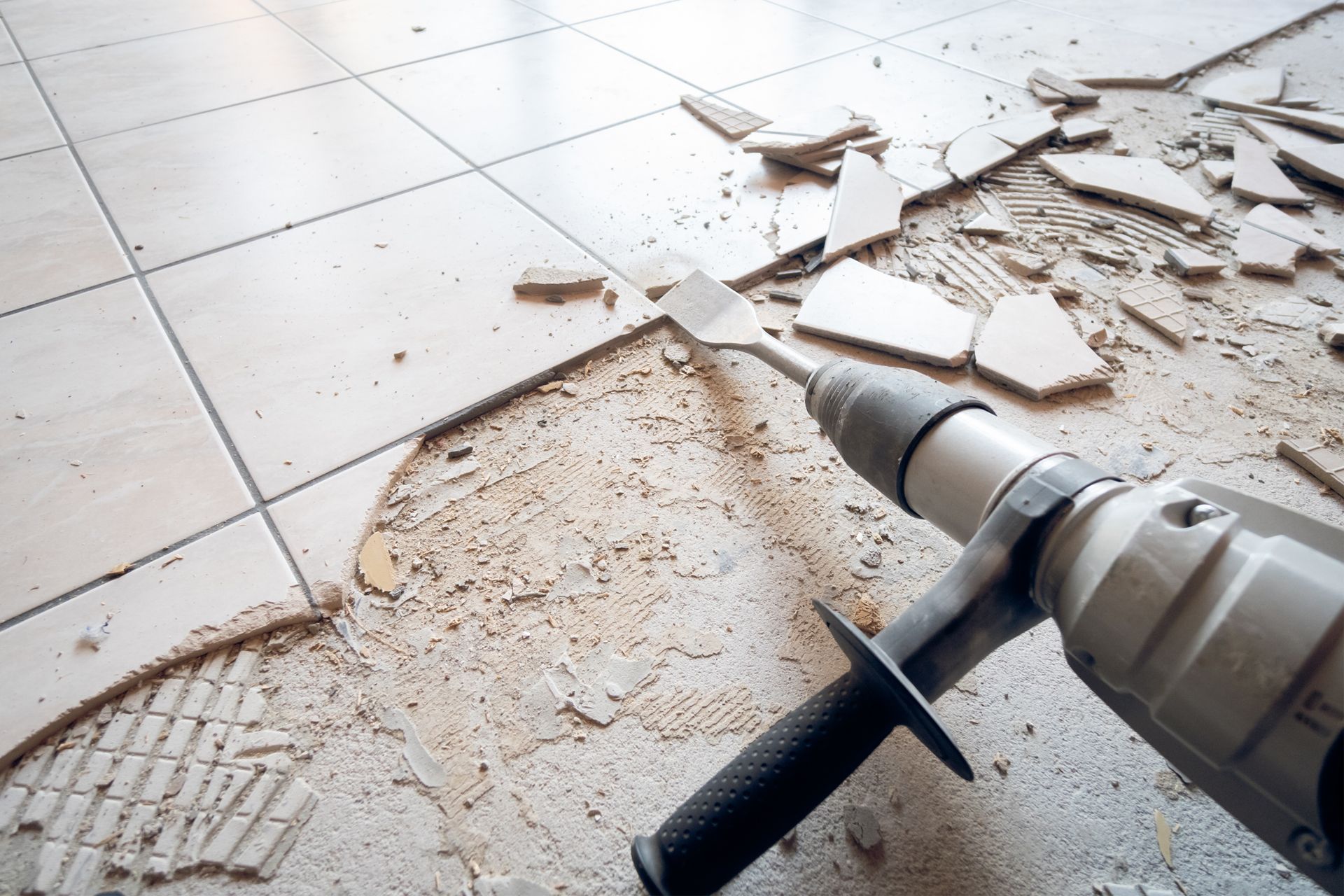 Tile floor being demolished with a drill and chisel bit, pieces of tile scattered.