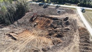 Aerial view of excavators digging trenches in a cleared, dirt-covered construction site near a road and trees.