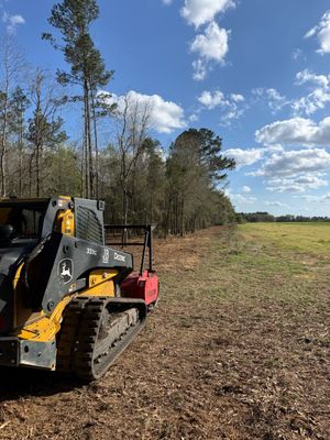 A yellow John Deere skid steer with a forestry mulcher attachment parked on cleared land next to a wooded tree line.