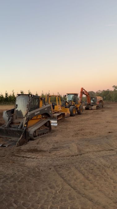 A row of heavy construction machinery, including bulldozers and an excavator, parked on a dirt lot at sunset.