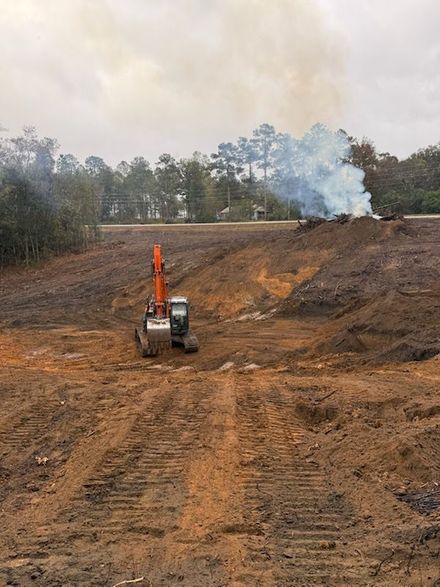 An orange excavator sits in a cleared, muddy construction site with a plume of white smoke rising in the background.