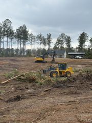 Two yellow pieces of heavy construction equipment clear debris from a wooded lot under a cloudy sky.