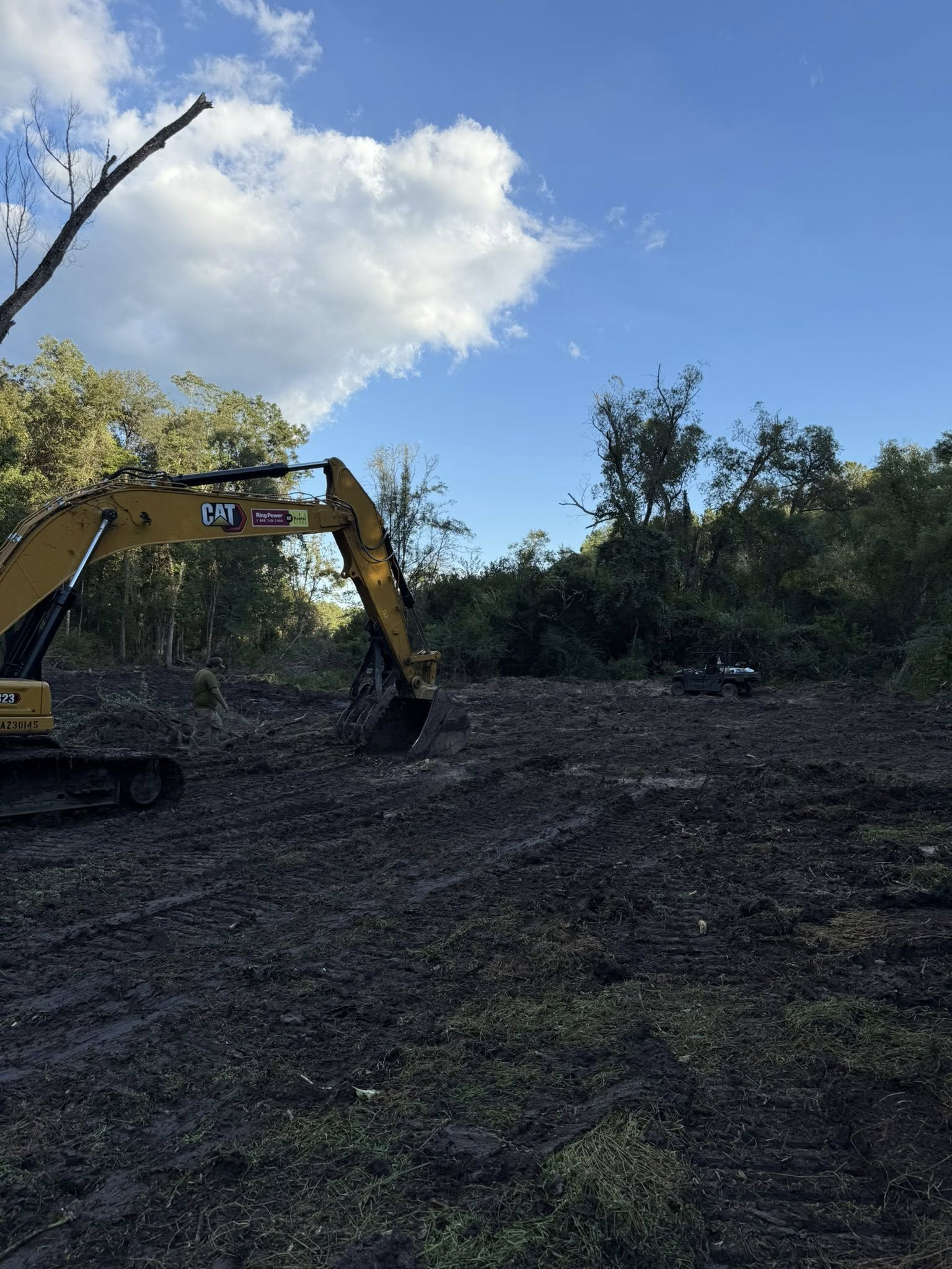 A yellow excavator sits in a muddy, cleared field with dense trees and a blue sky in the background.