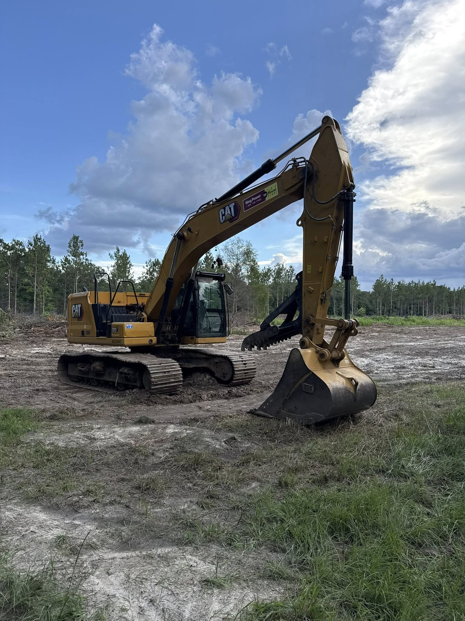 A yellow Caterpillar excavator sits on a cleared, earthy plot of land under a bright, cloudy sky.