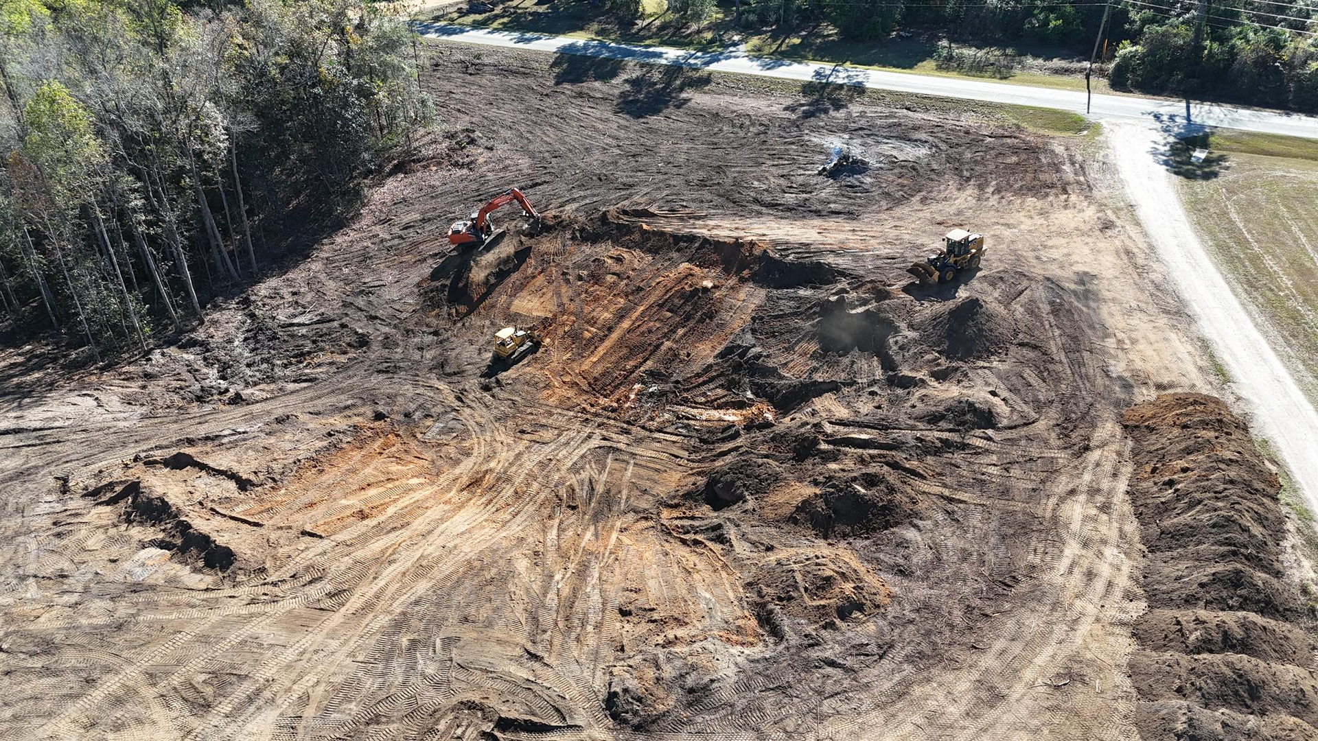 An aerial view of an active construction site with machinery excavating soil in a cleared, wooded area near a road.