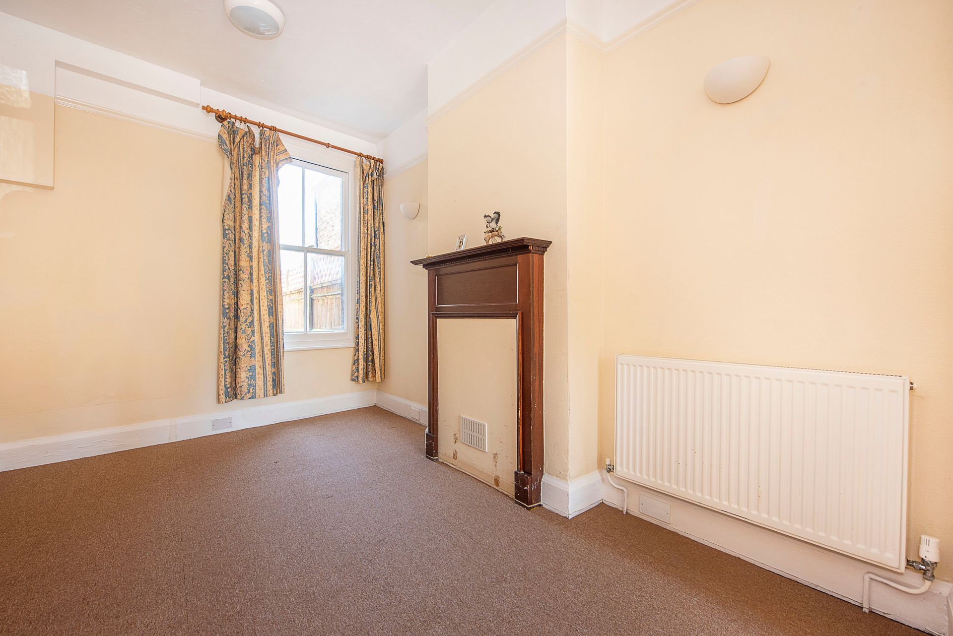 Empty room with brown carpet, cream walls, fireplace, window, and radiator.