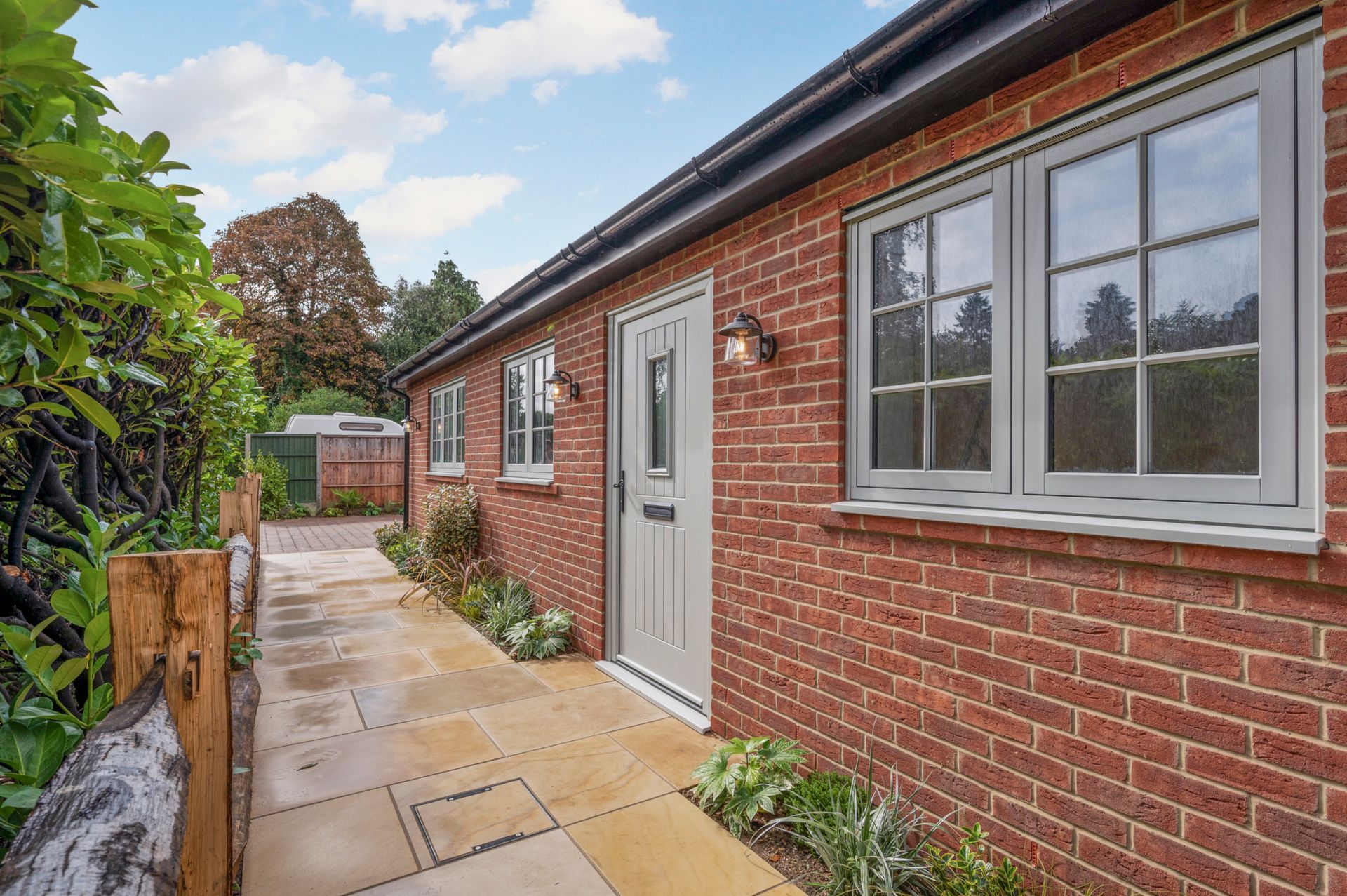 Brick building with gray windows and door, walkway, plants, and greenery under a partly cloudy sky.