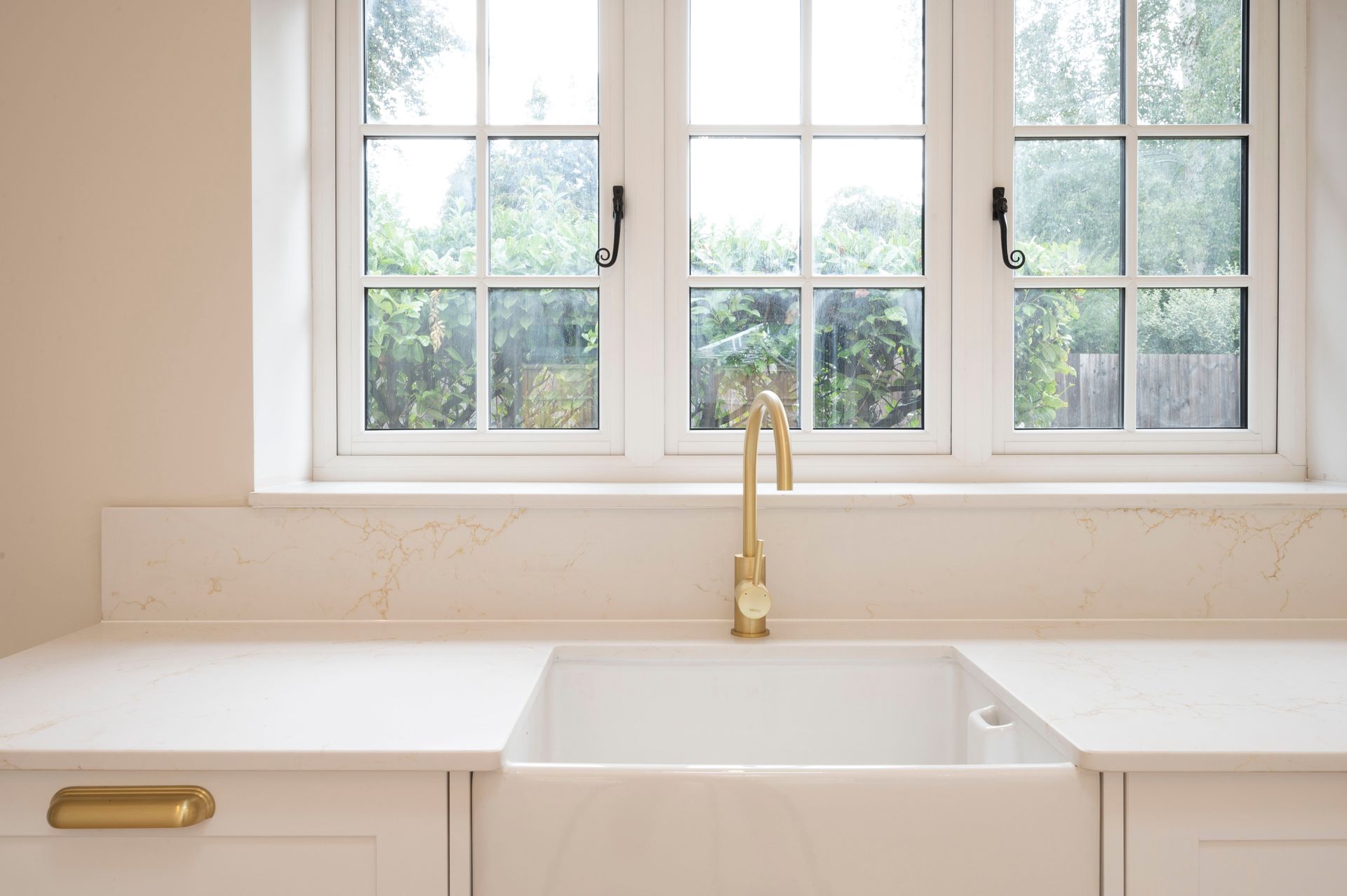 White kitchen sink with gold faucet under a grid window, creamy countertops, and a white cabinet.