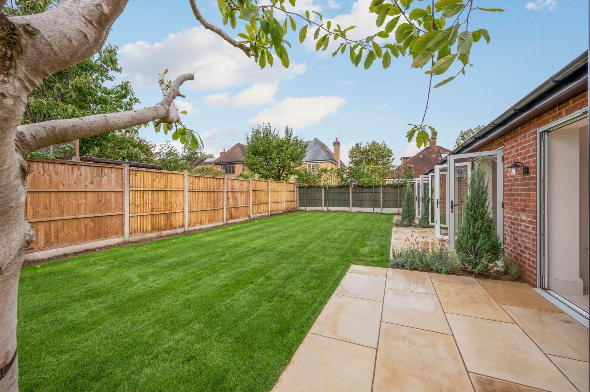 Green lawn and patio in a backyard, wooden fence, brick house, blue sky.
