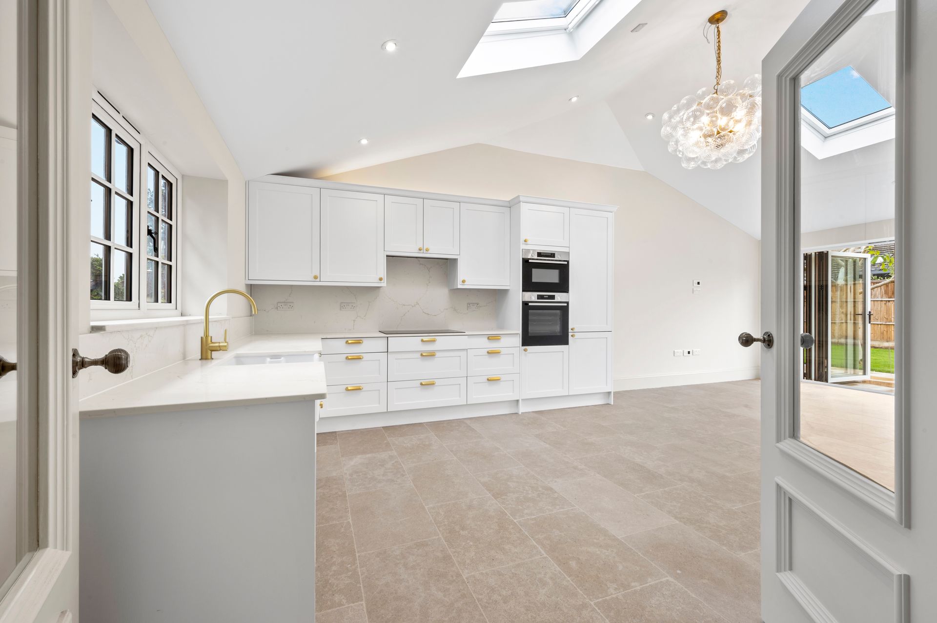 Bright, white kitchen with skylights, white cabinets, gold fixtures, and a view of a backyard.