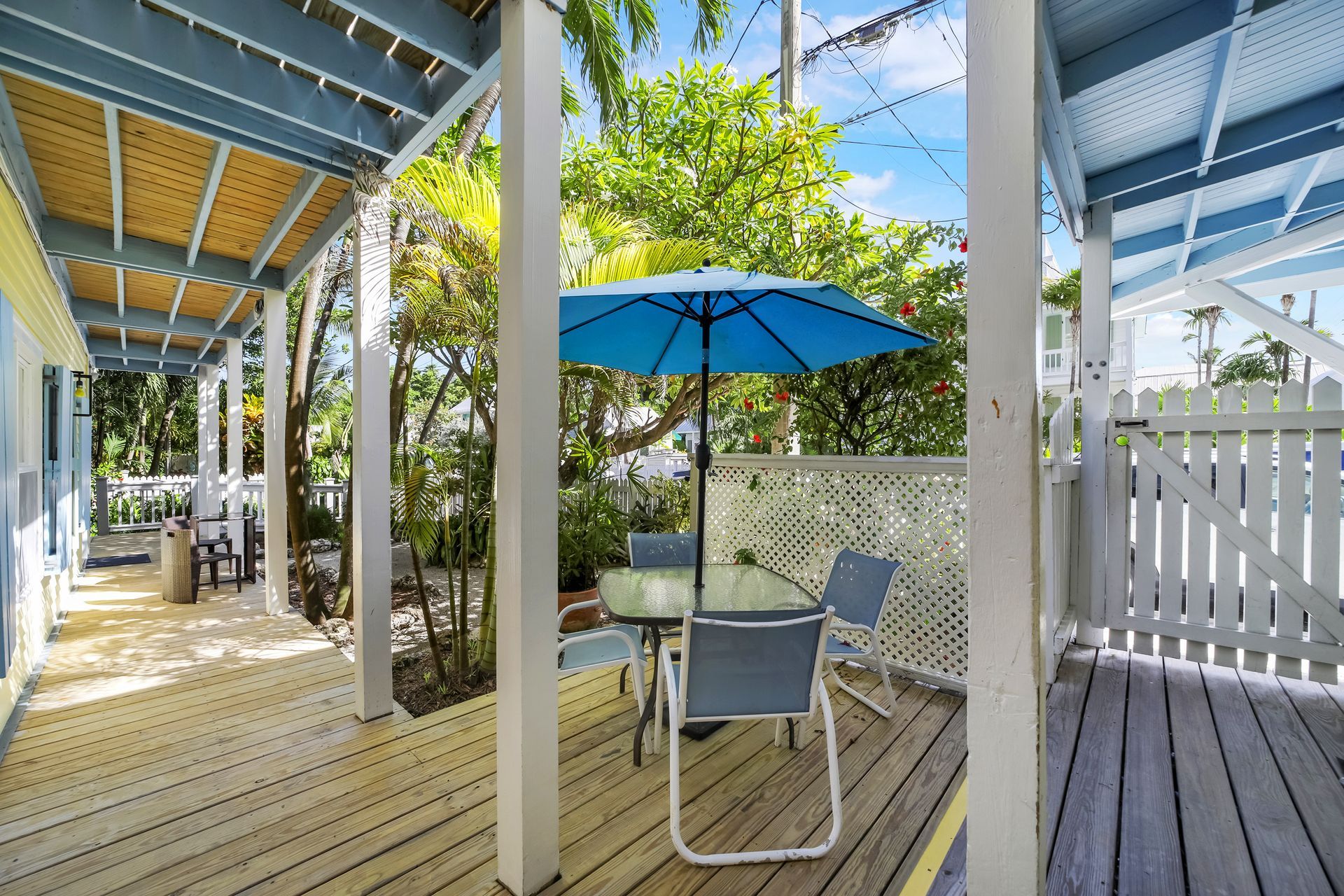 Wooden porch with table, chairs, and blue umbrella; trees and sky in the background.