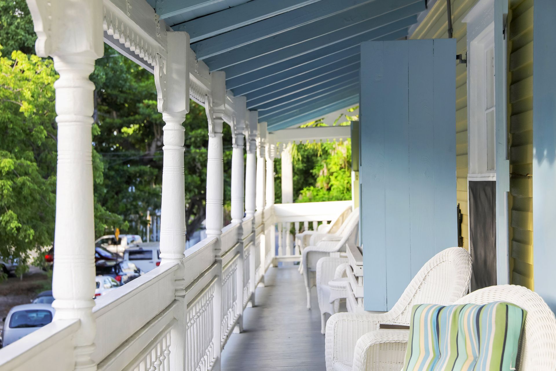 White porch with ornate columns, blue ceiling and door, overlooking a street with parked cars and trees.