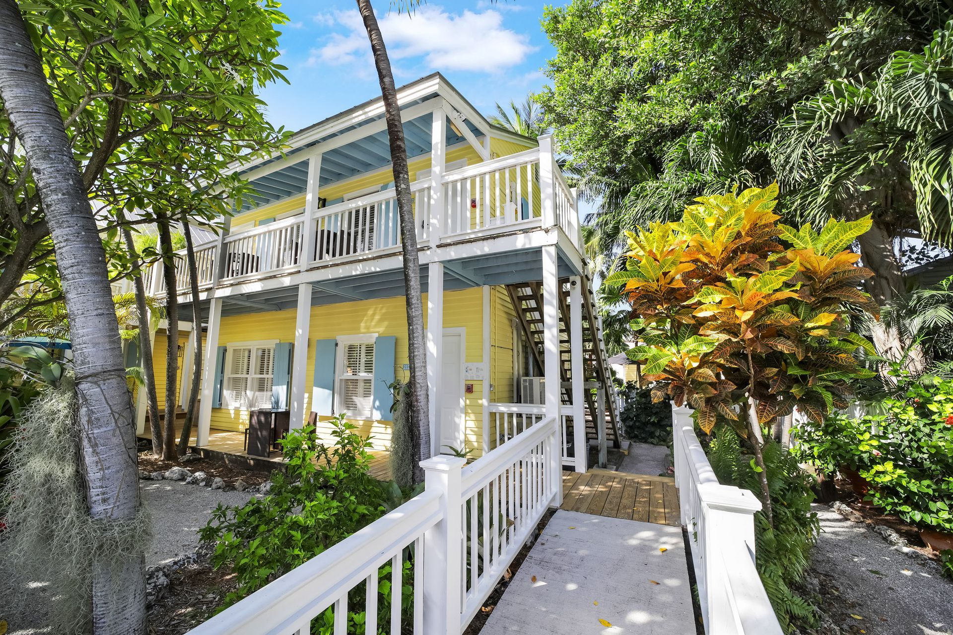 Two-story yellow house with white balconies, blue shutters, and tropical foliage.