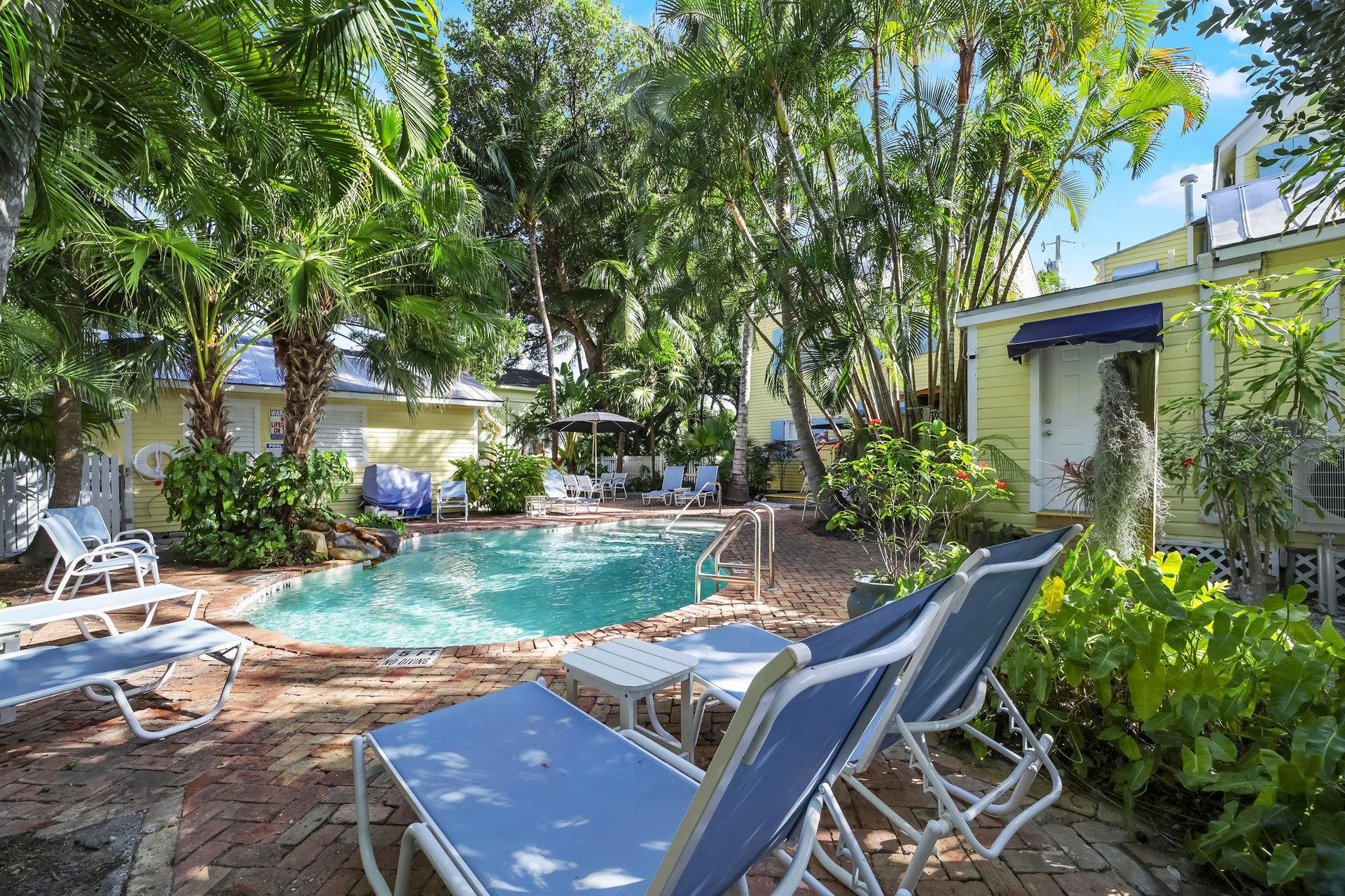 Pool surrounded by trees, lounge chairs, and a yellow building under a sunny sky.