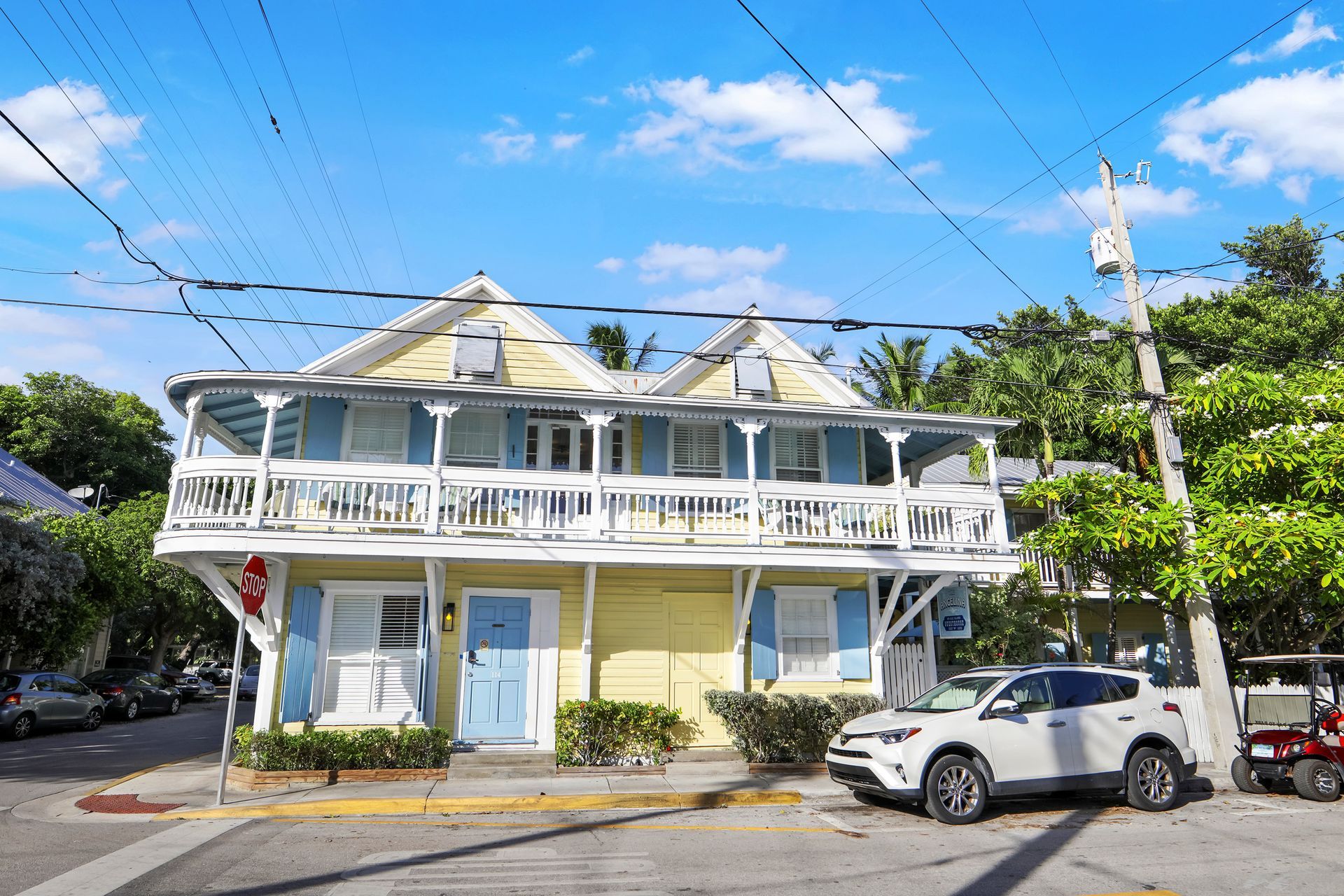Two-story yellow house with blue shutters and a balcony in Key West, Florida. A white SUV is parked in front.