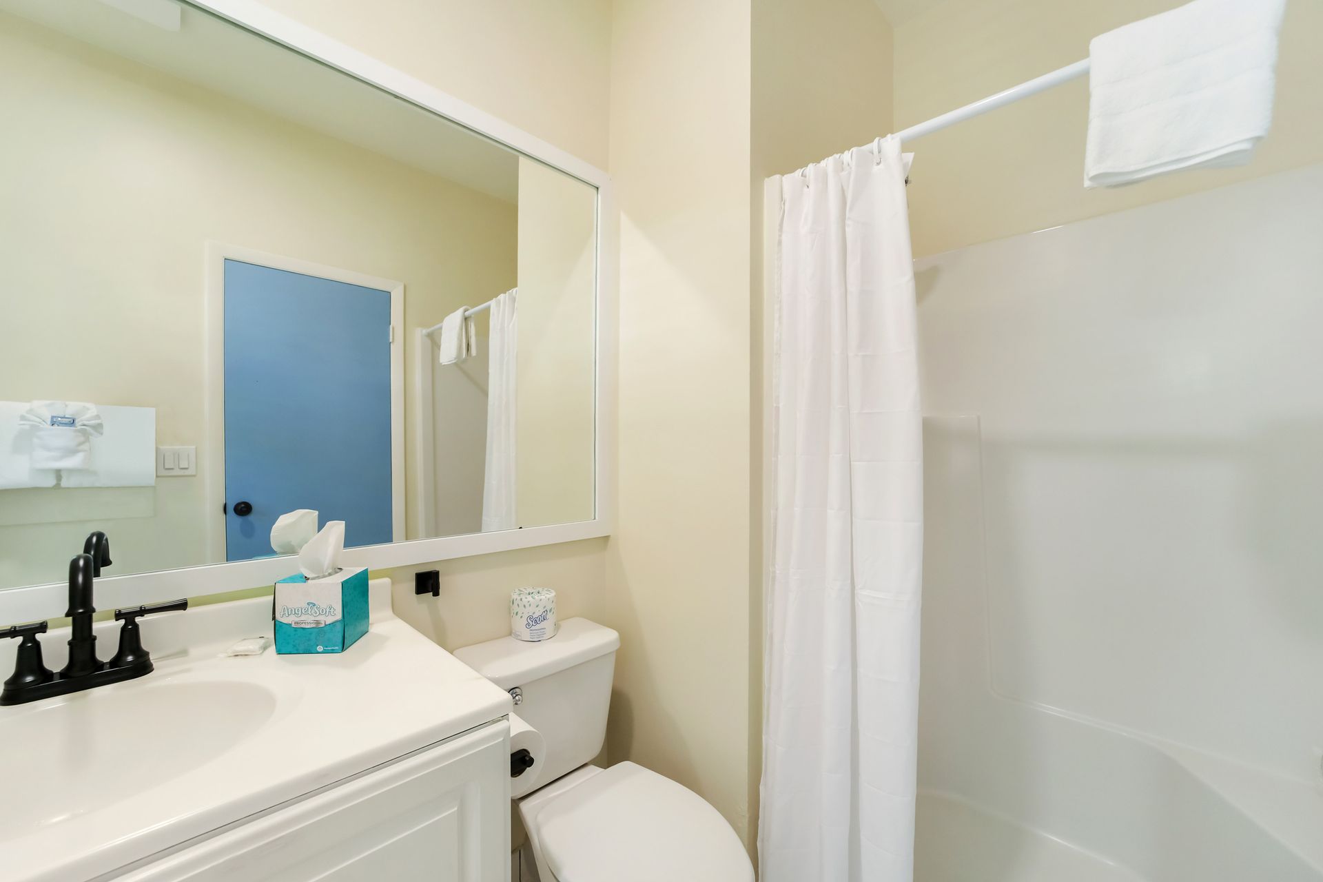 Bathroom with white sink, toilet, and shower/tub. Blue door in the mirror. Light beige walls.