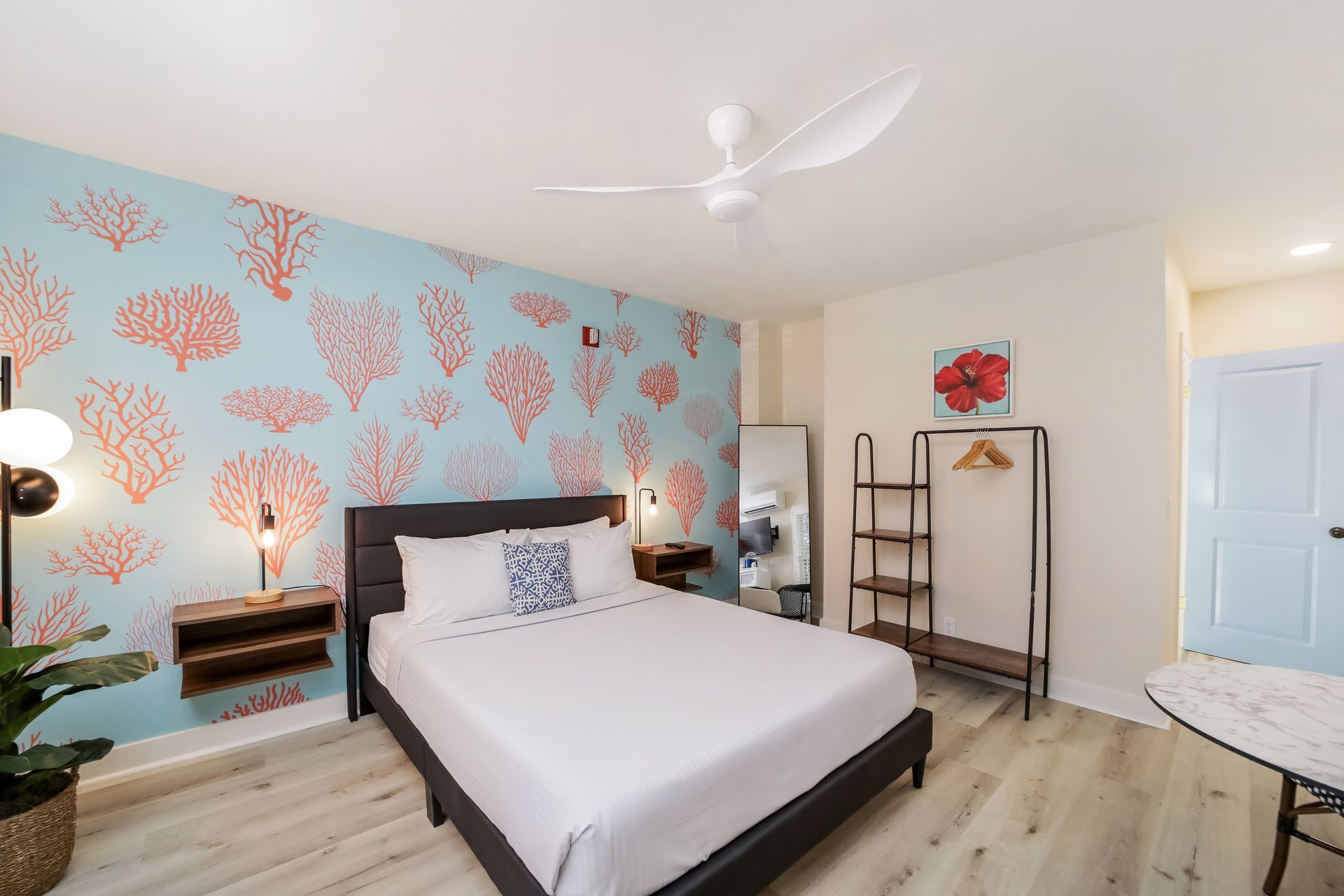 Bedroom with coral wallpaper, a bed, a mirror, and a table. Light wood floor and a ceiling fan.