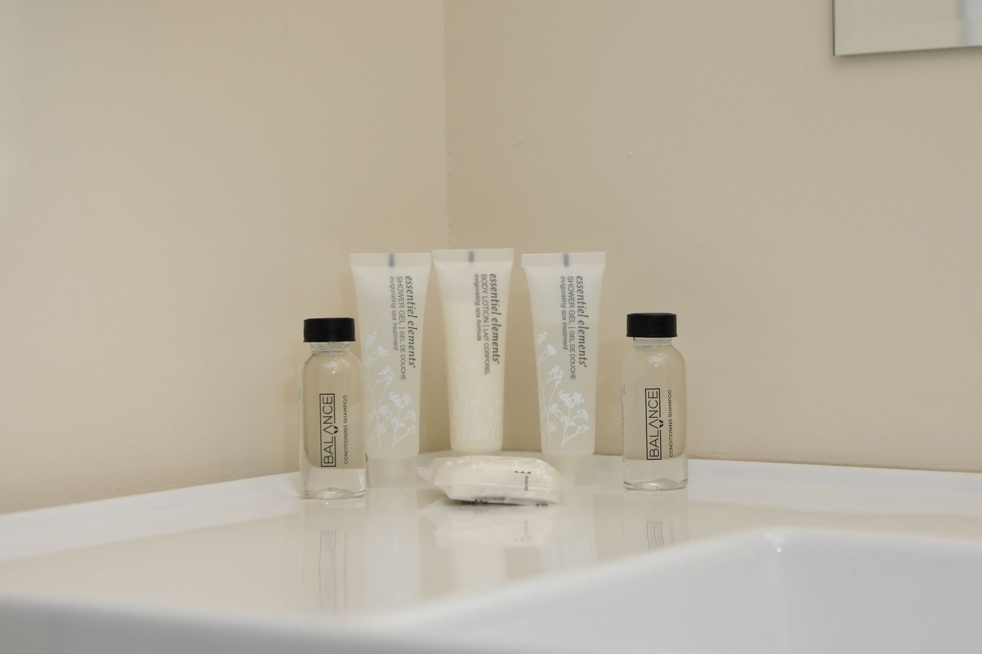 Bathroom toiletries on a white sink: shampoo, conditioner, lotion, and soap, against a beige wall.