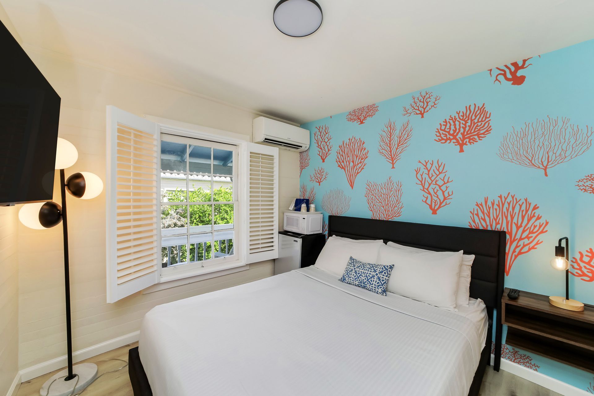 Bedroom with coral-themed wallpaper, a bed, a window with shutters, and a floor lamp.