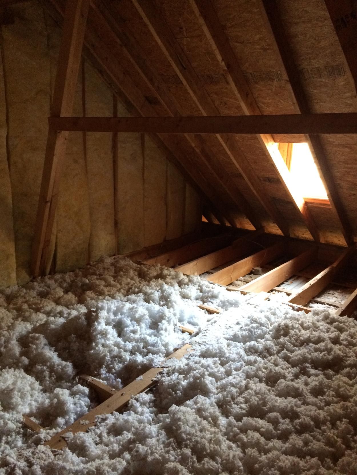 Interior view of an attic filled with white insulation, wooden beams and a small window letting in sunlight.