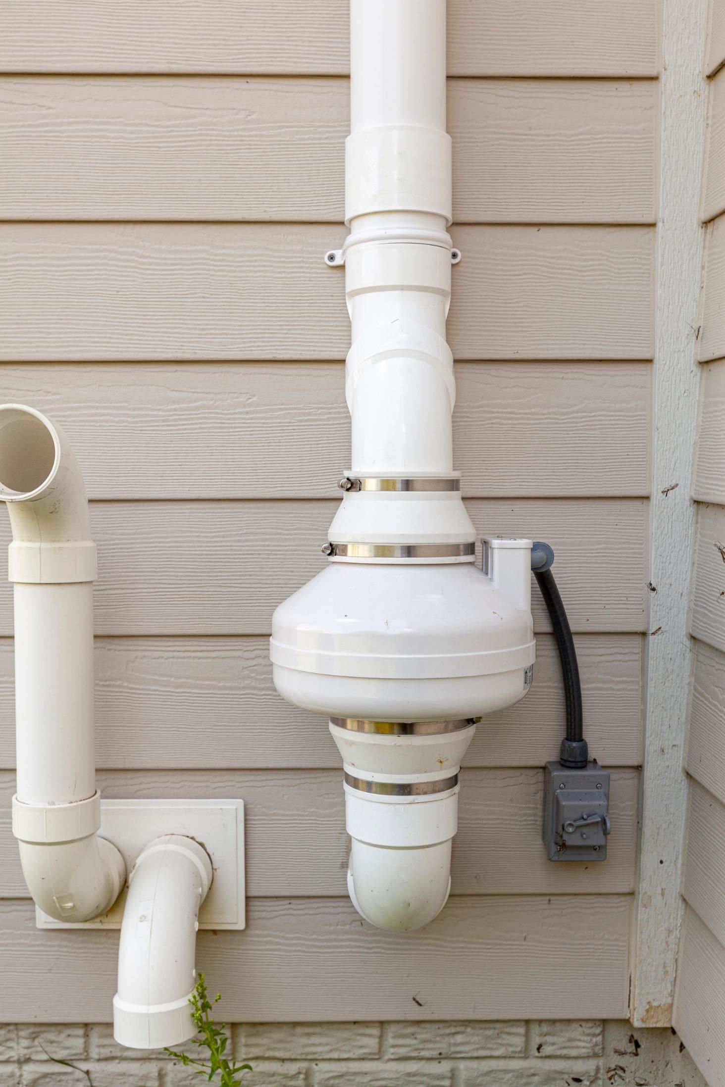 White radon mitigation system on a light brown siding, with a power outlet on the right.