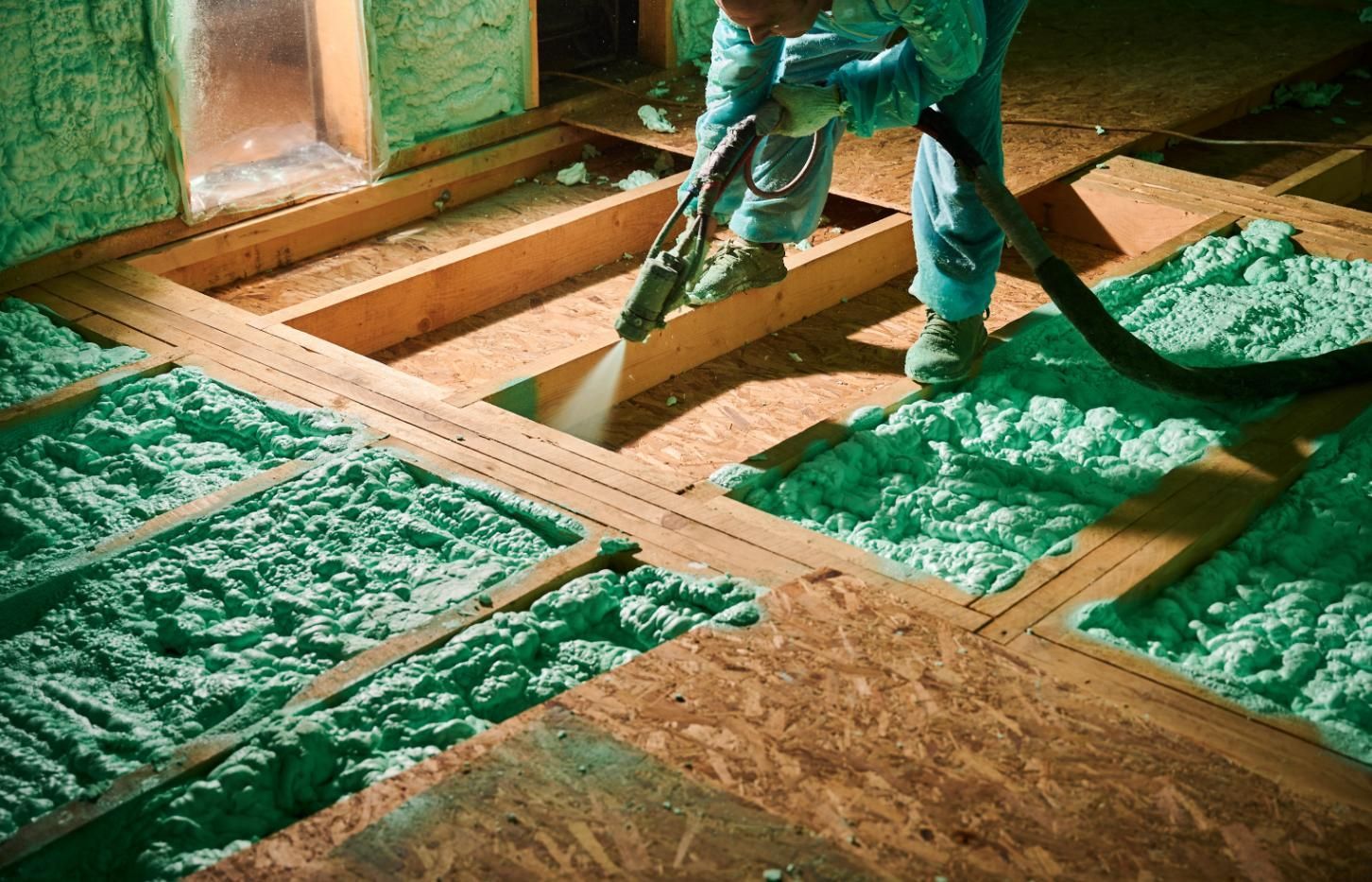 Person spraying green foam insulation into a floor's wooden frame, in an unfinished room.