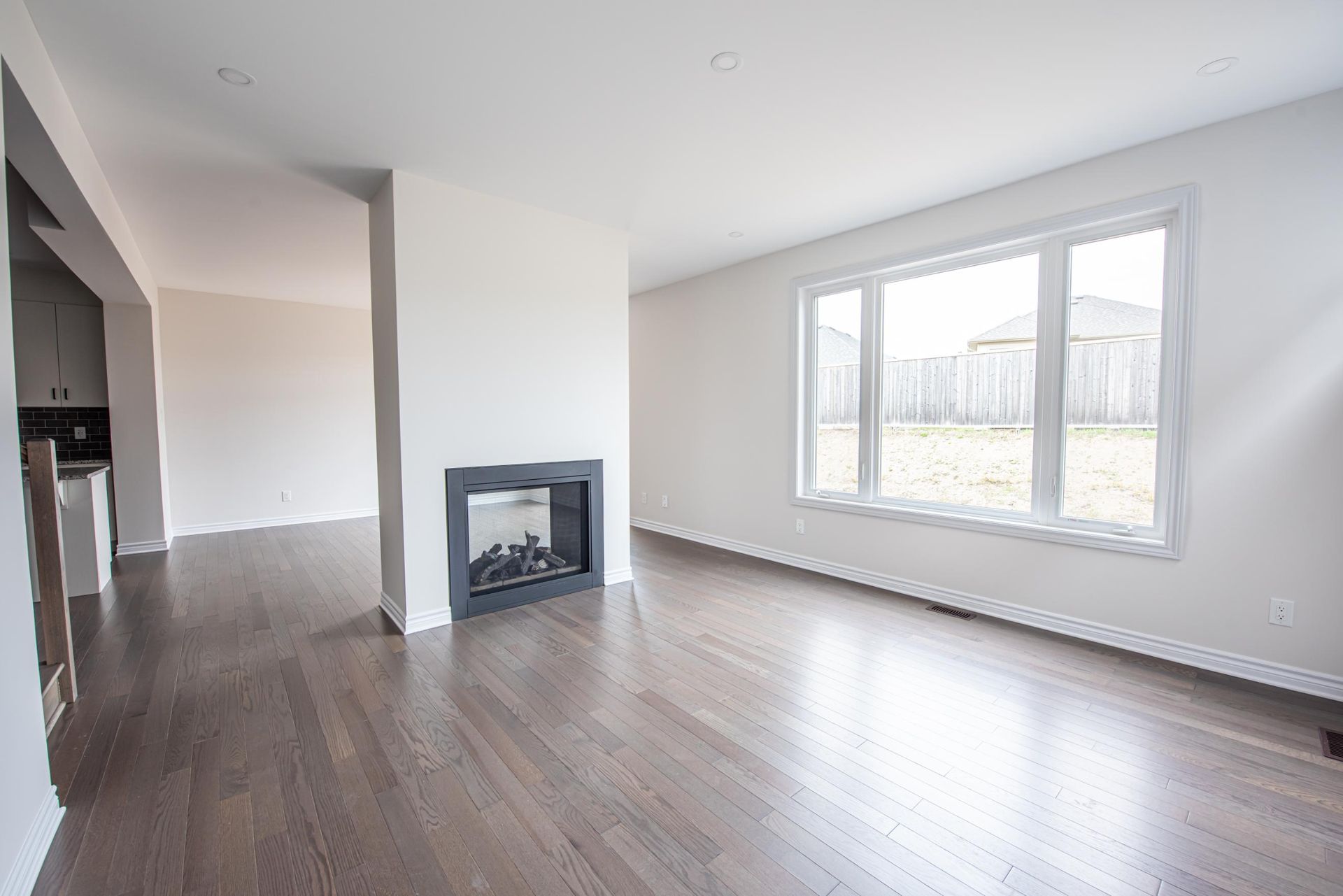 Empty living room with dark wood floors, fireplace, and window overlooking a backyard.