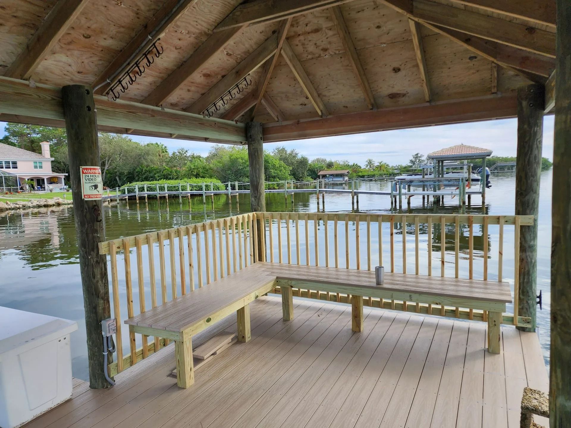 A covered wooden dock with two benches overlooking a body of water, a pier, and houses in the background.