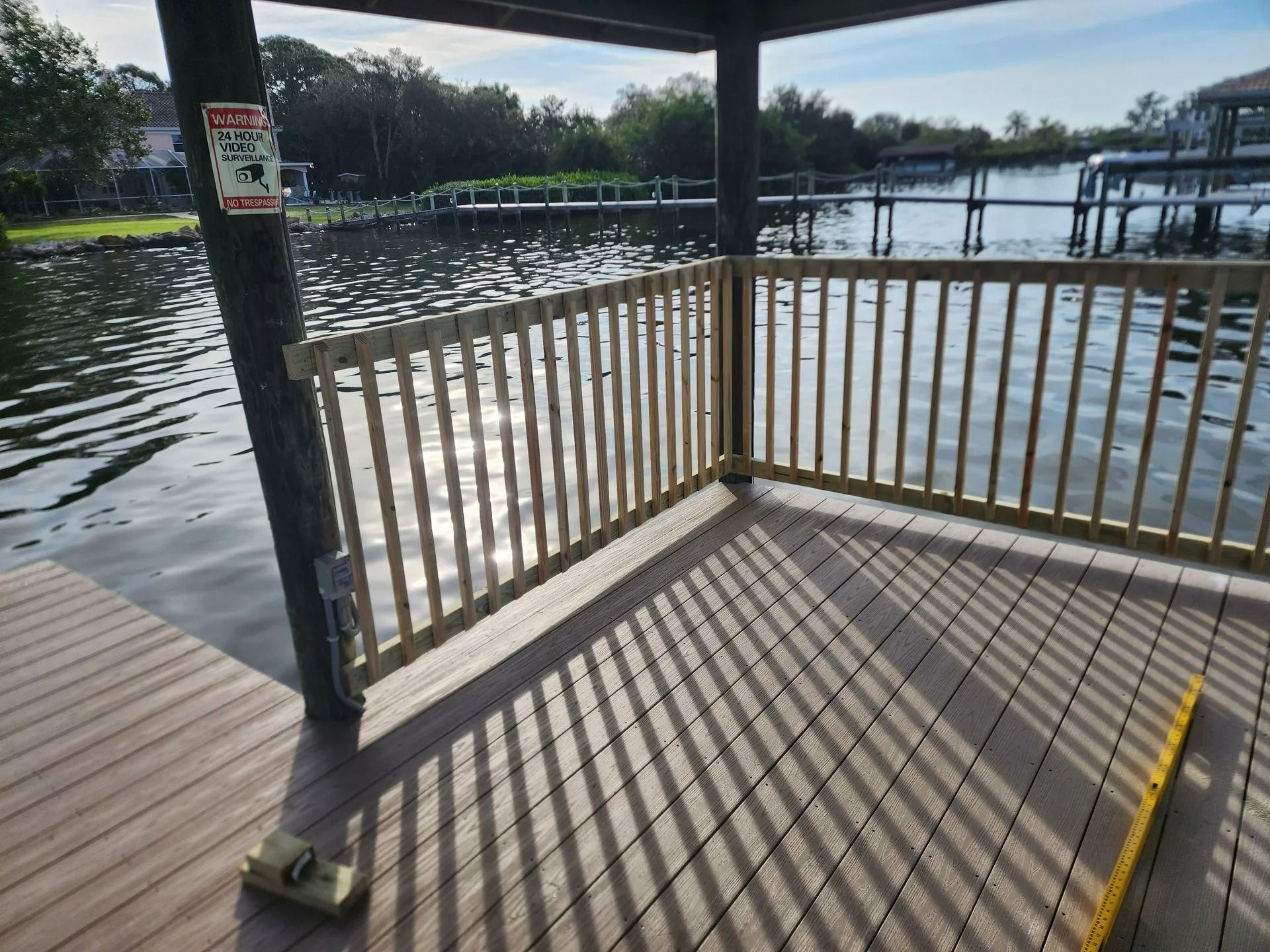 A wooden dock with a railing overlooking water. Sunlight casts shadows on the deck.