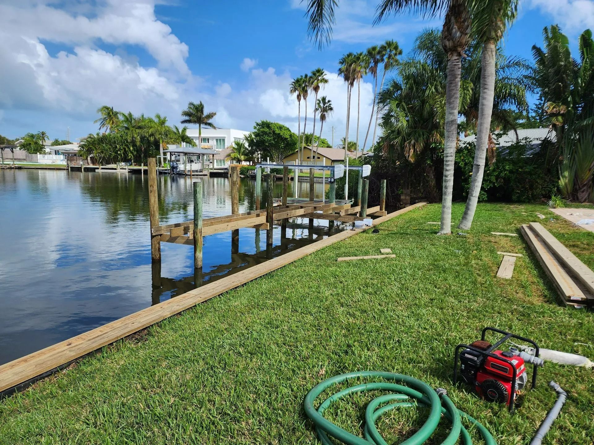Dock on a canal, with palm trees and homes in the background. Green grass and a water hose are in the foreground.