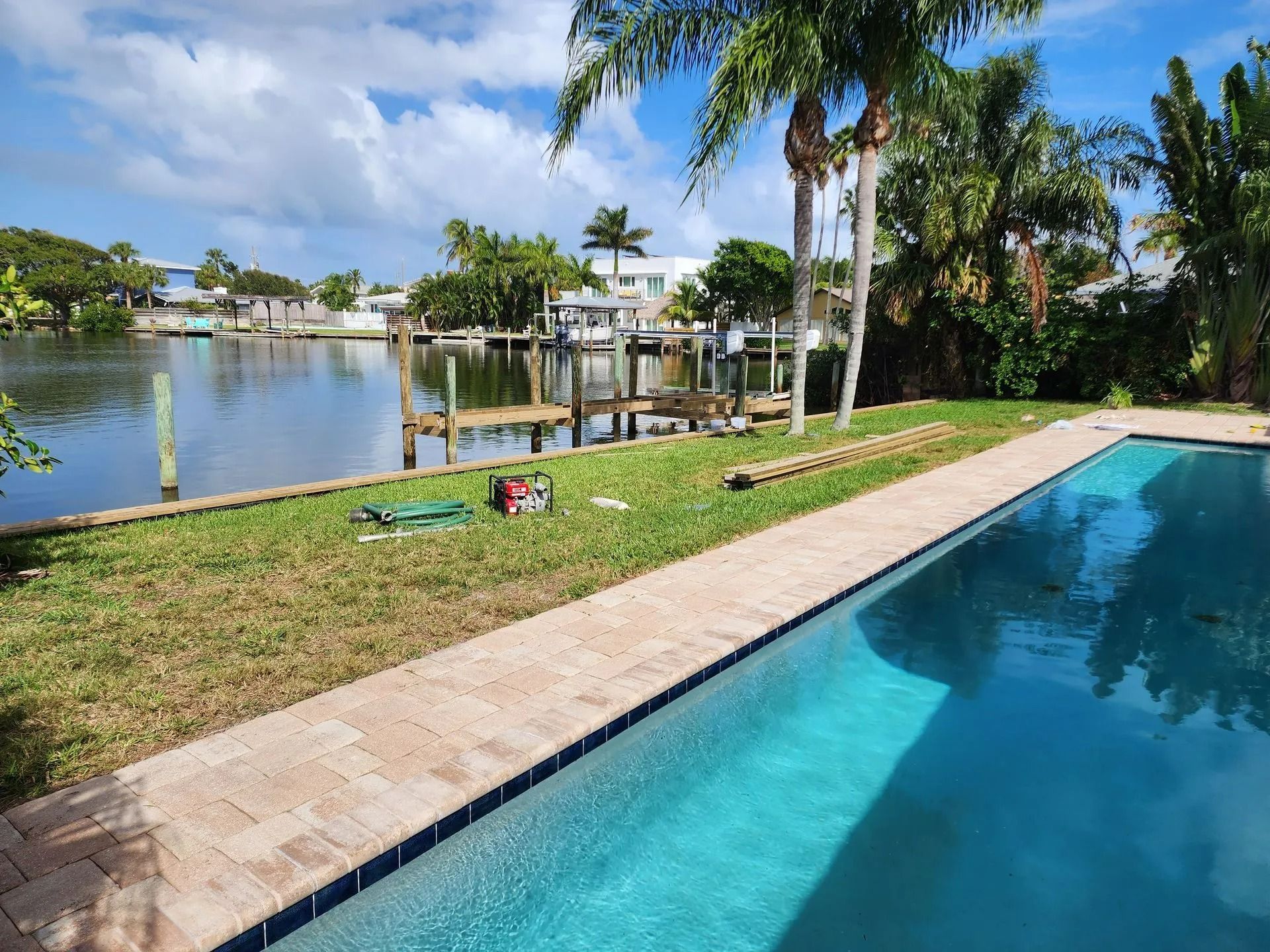 A swimming pool next to a canal.  A dock and houses are visible across the water. Green grass and palm trees surround the pool.