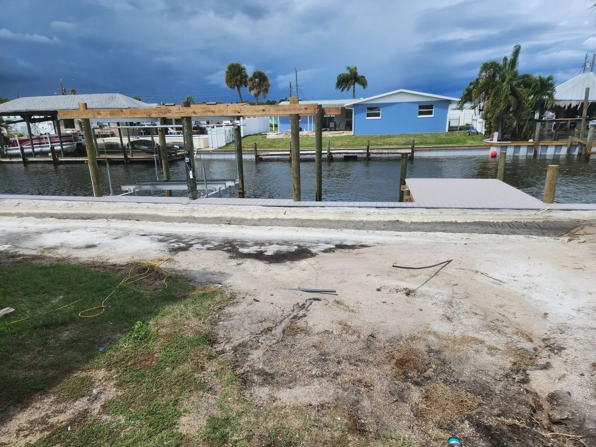 Canal with wooden dock supports, unfinished construction, and a light blue house under a cloudy sky.