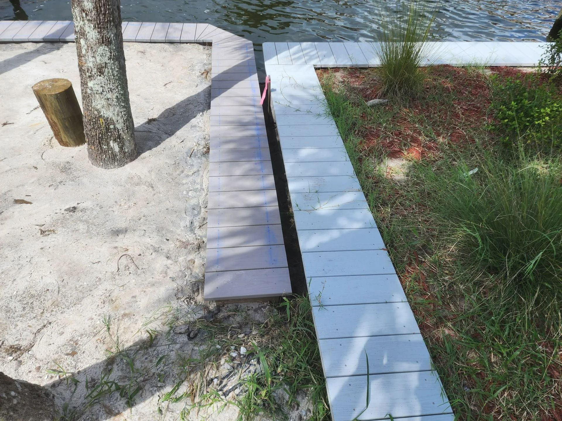 A wooden boardwalk extends along a sandy beach and grassy area, next to a body of water and a tree.