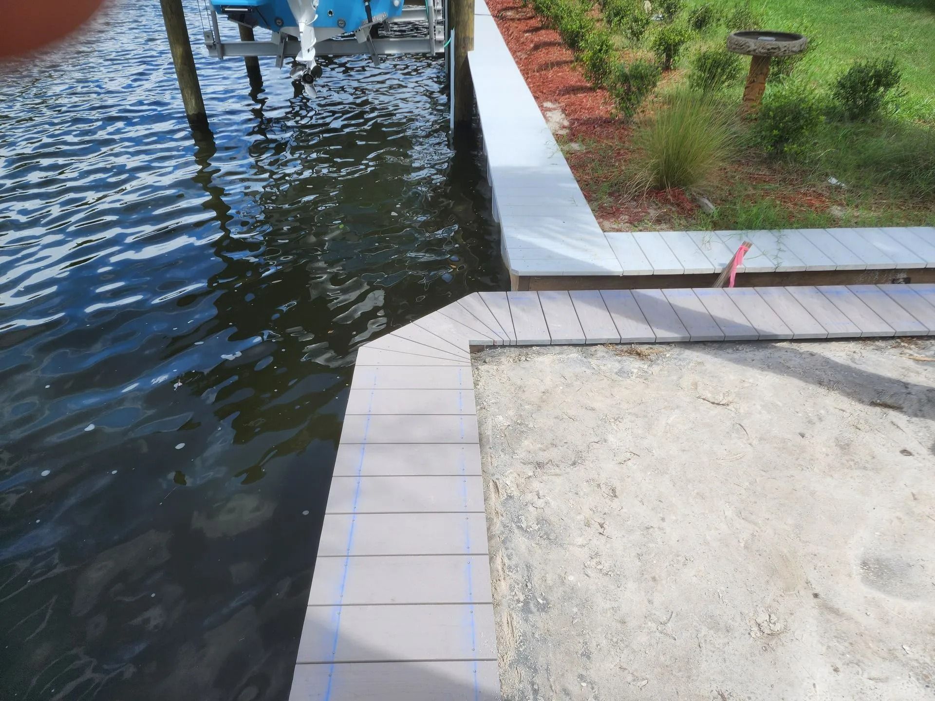 A waterfront view of a composite deck edging a sandy area. Water and a boat are visible to the left. Landscaping is to the right.