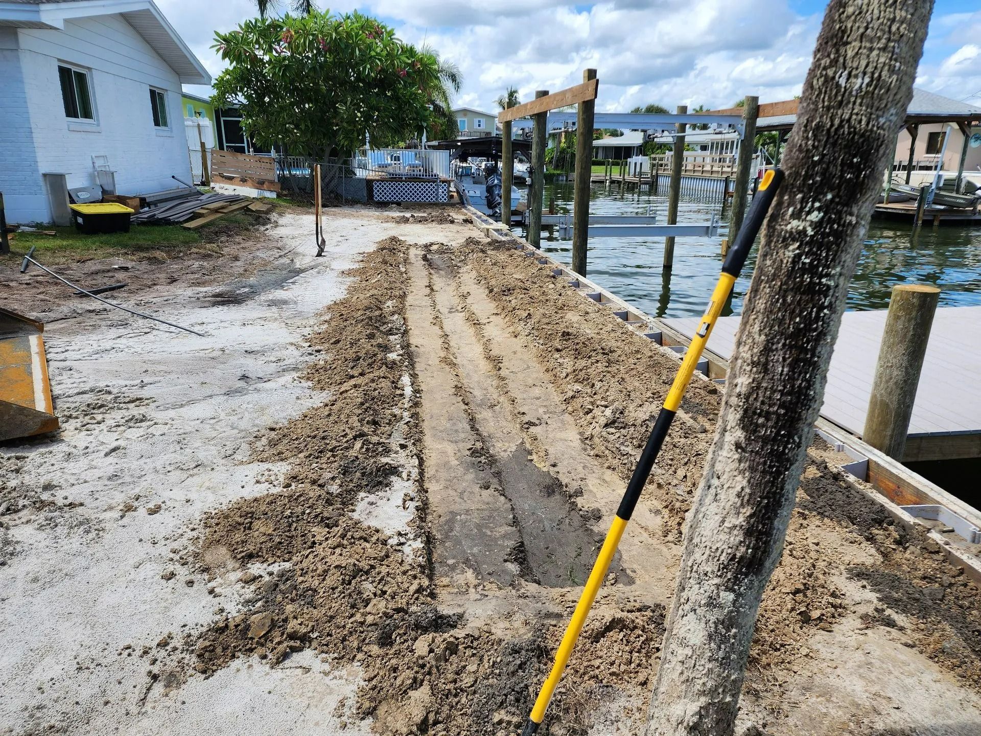 A dock is under construction with a trench dug along the concrete surface; a shovel leans against a wooden post.
