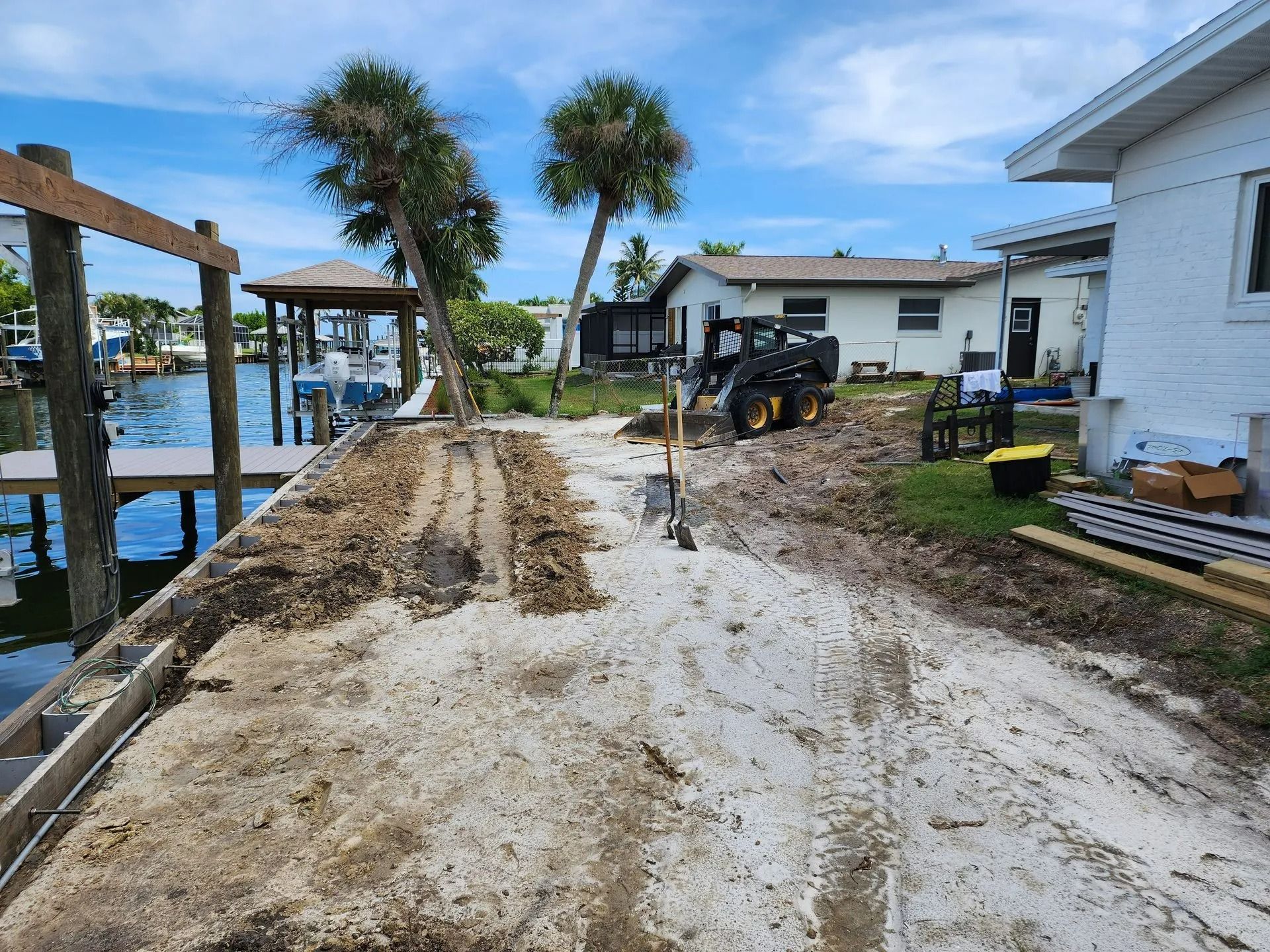 Dirt and sand path along a waterfront with construction underway. A backhoe, dock, and houses are in the background under a blue sky.