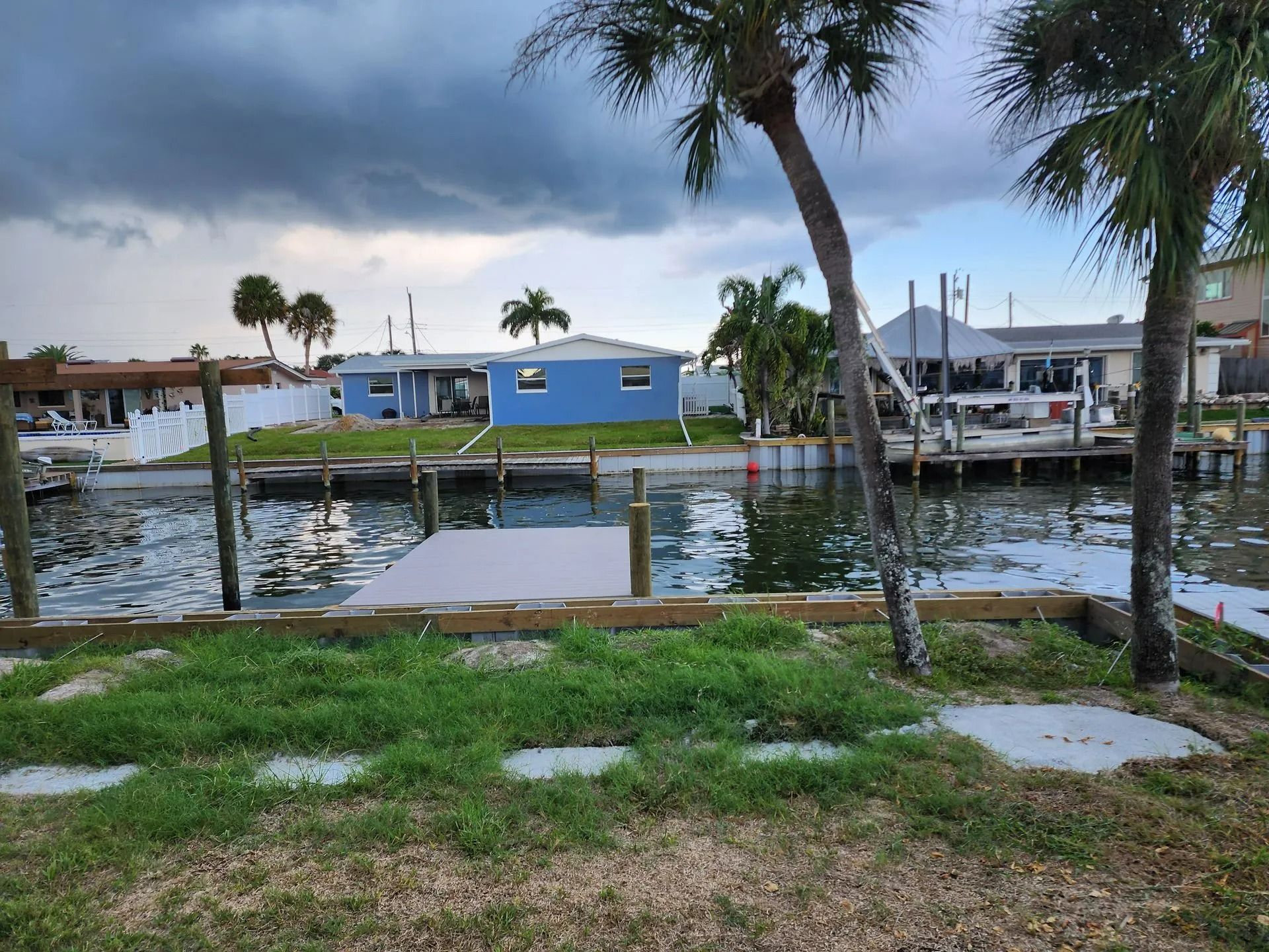 Coastal homes along a canal under a cloudy sky, with docks and palm trees in the foreground. A blue house is centered.