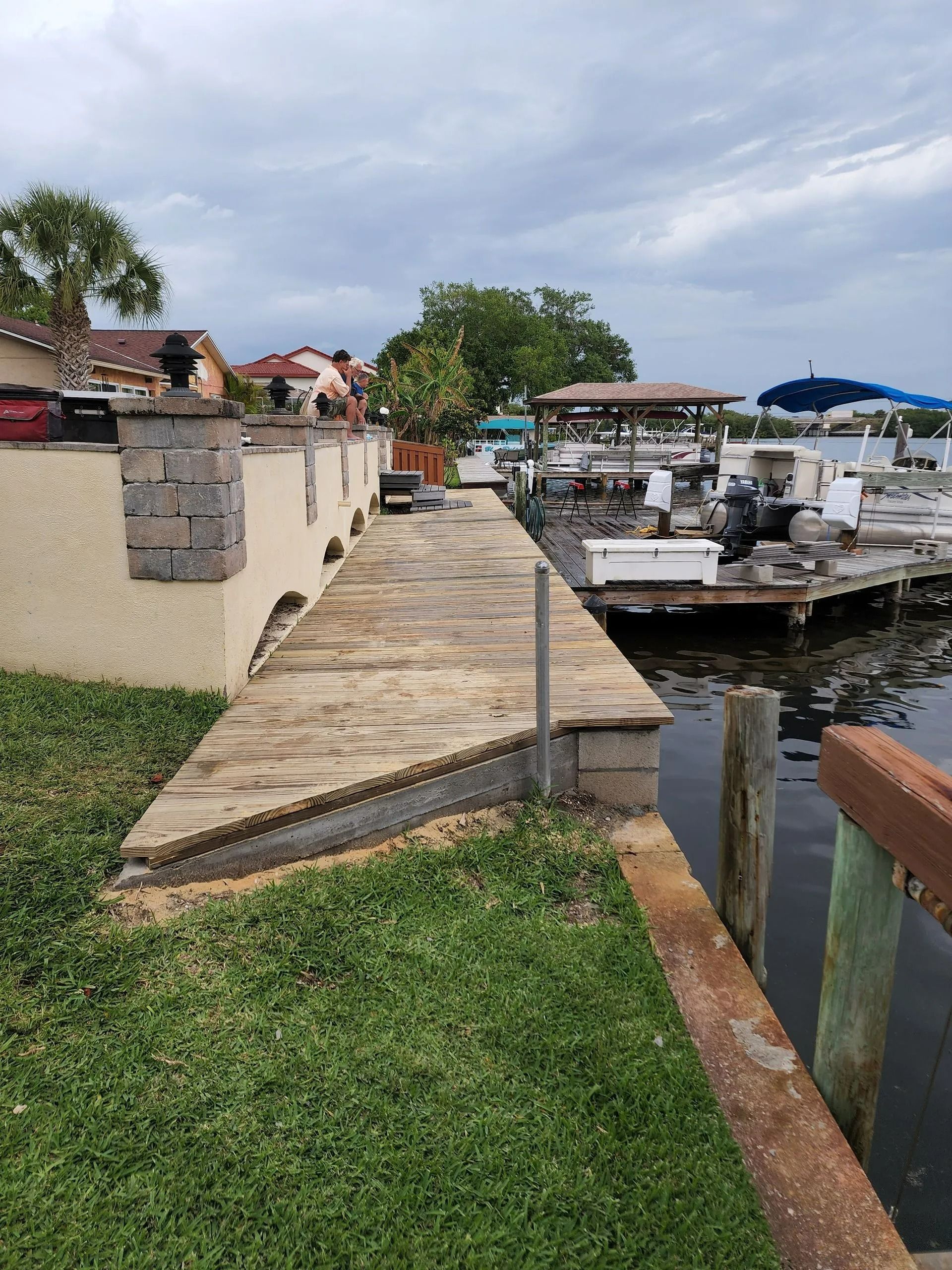 Brick walkway along a waterfront, leading towards a dock with boats under a cloudy sky. Green grass borders the walkway.