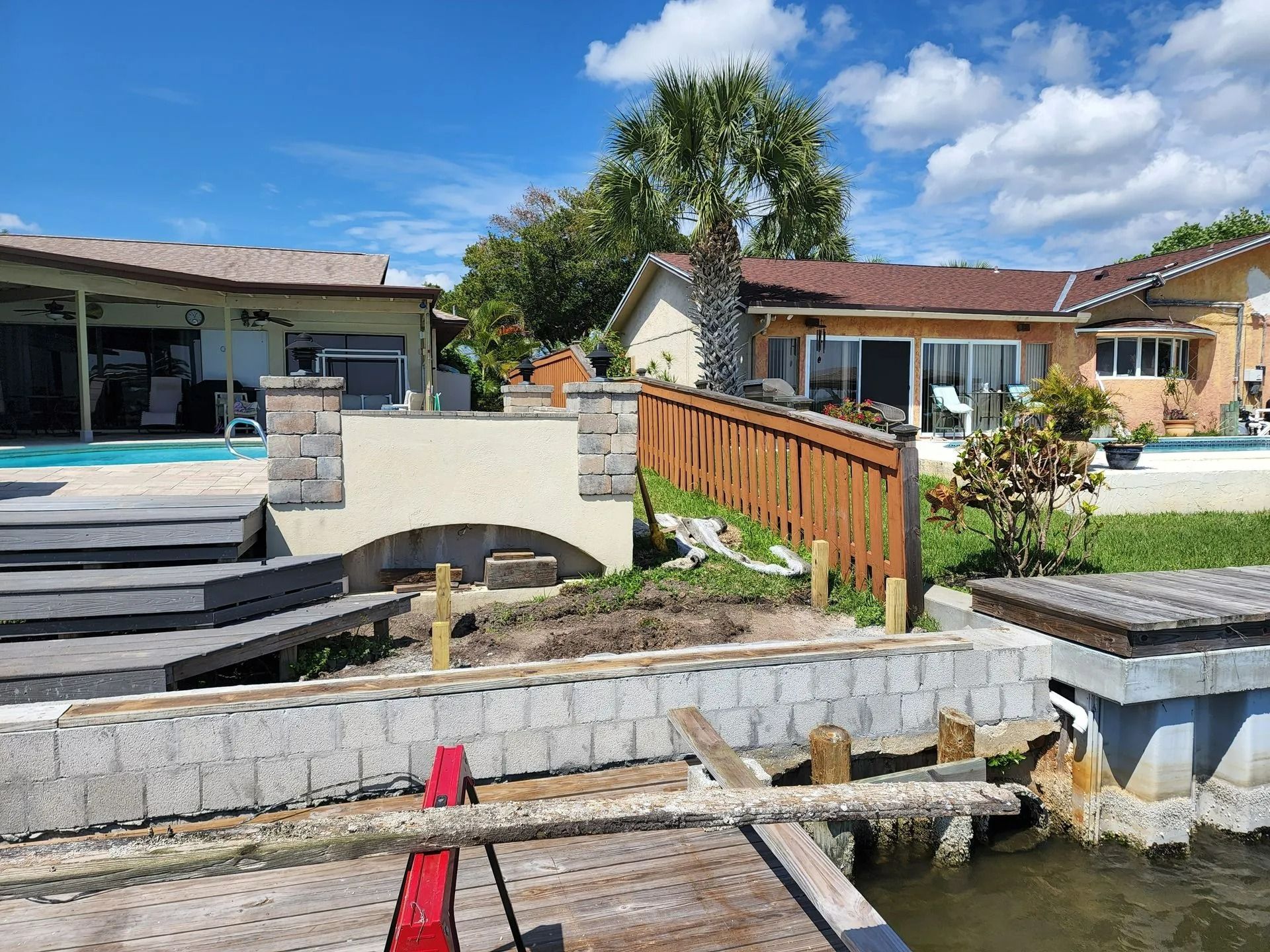 A waterfront home with a pool, bridge, and dock. A wooden fence and stone walls frame the scene on a sunny day.