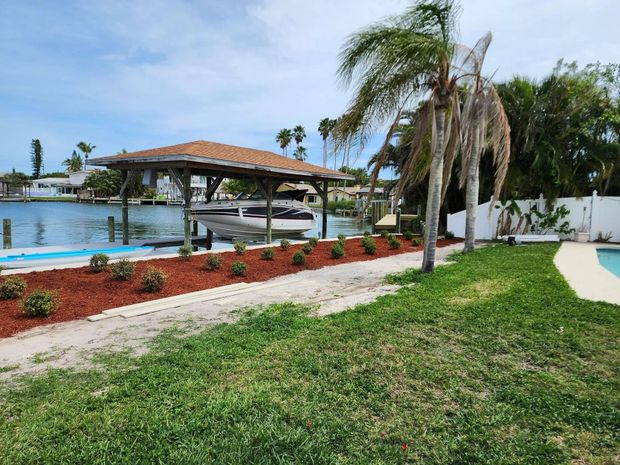 A boat dock with a covered boat lift sits on a canal. Red mulch borders a gravel path next to green grass and a white fence.