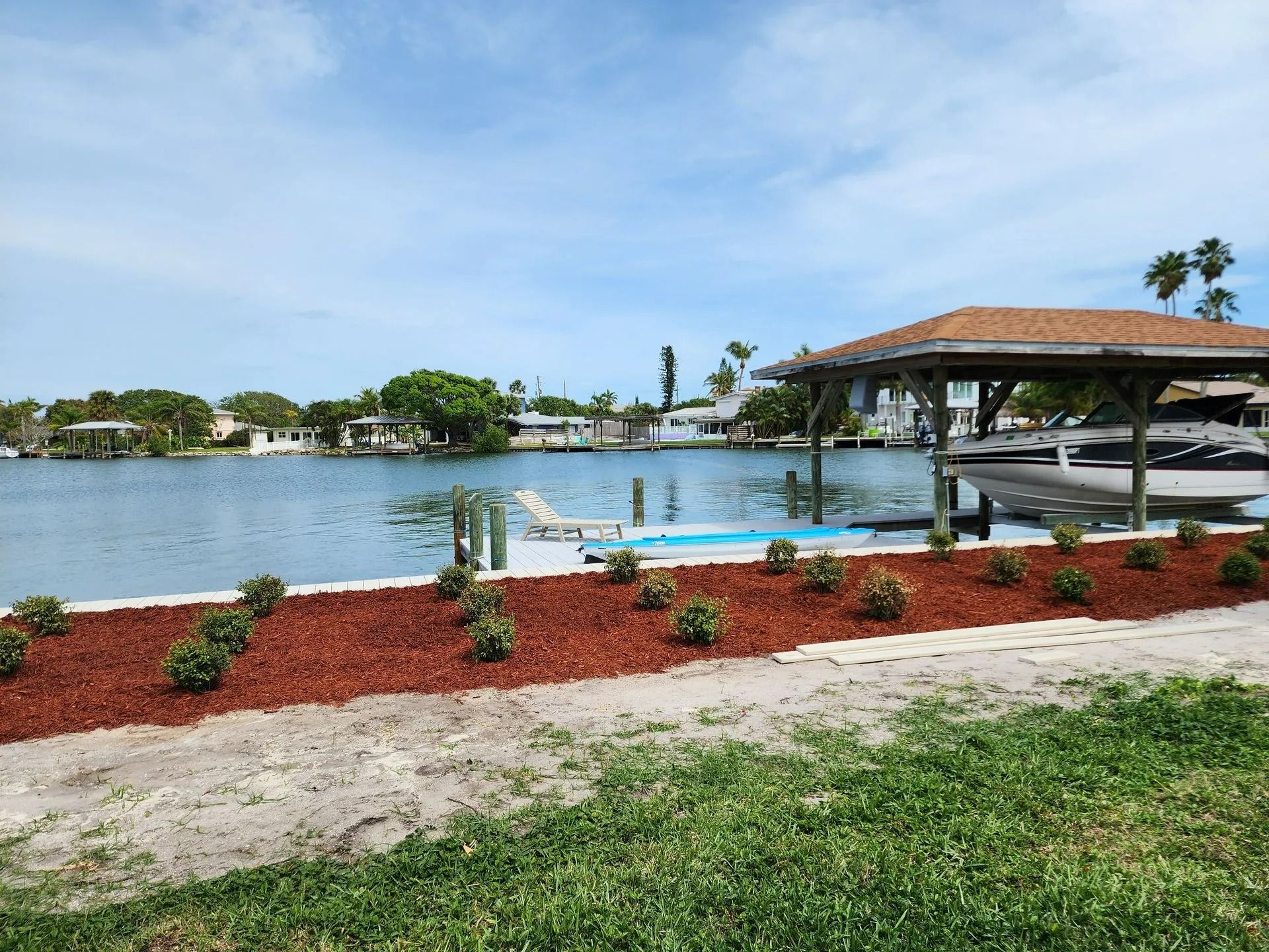 A waterfront view with a dock, shrubs with red mulch, and houses on the water under a blue sky.
